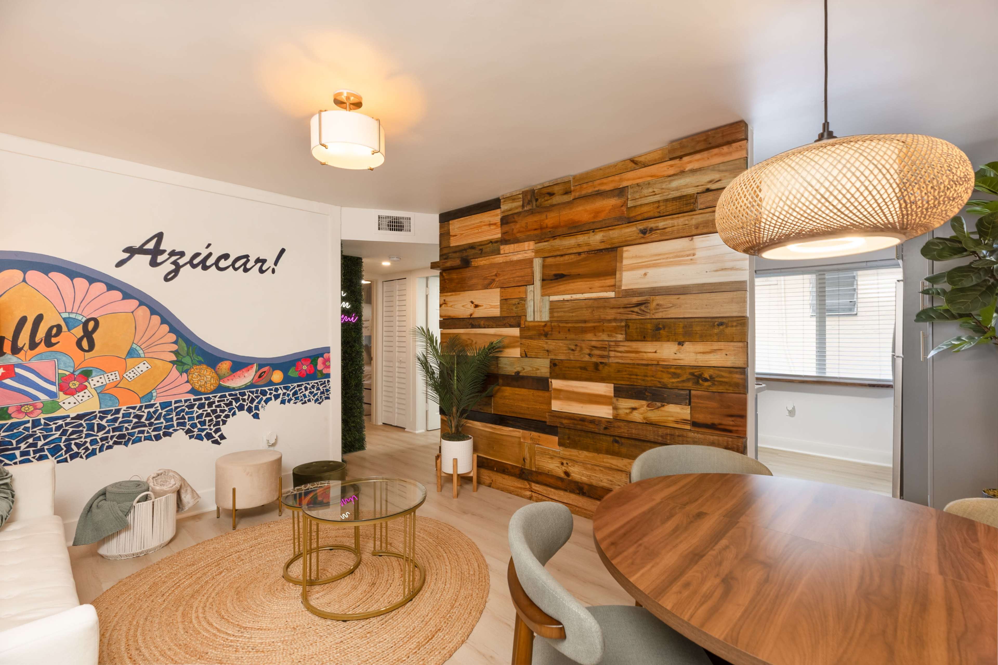 The image shows a modern living space featuring a dining area with a round wooden table, a decorative wall with colorful artwork, and a textured wooden accent wall.