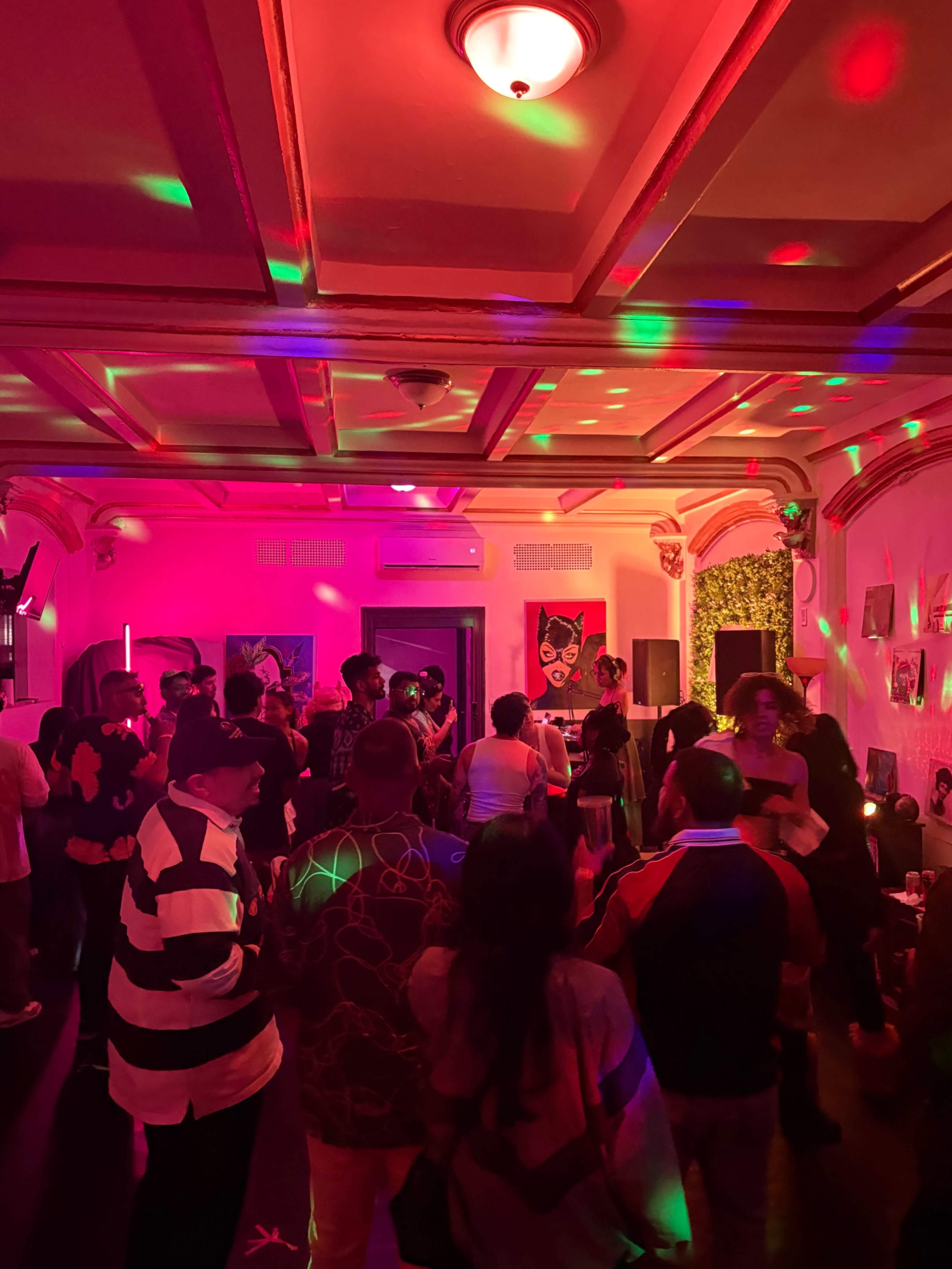 A crowded room features people socializing and dancing under colorful lights, with artwork displayed on the walls.