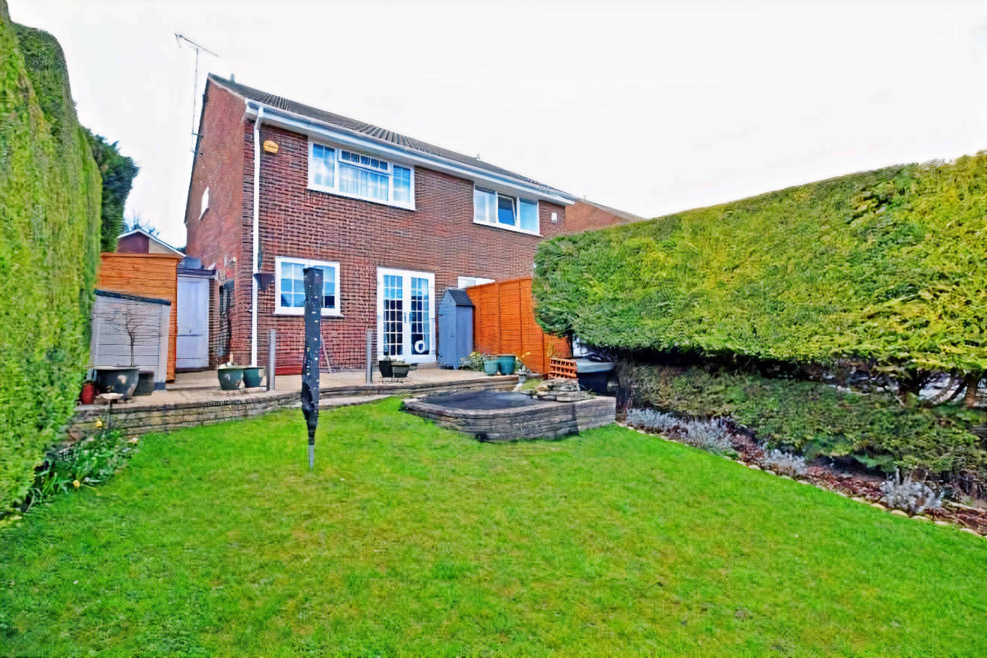 The image shows a two-story brick house surrounded by neatly trimmed hedges and a small garden area with stone pathways.