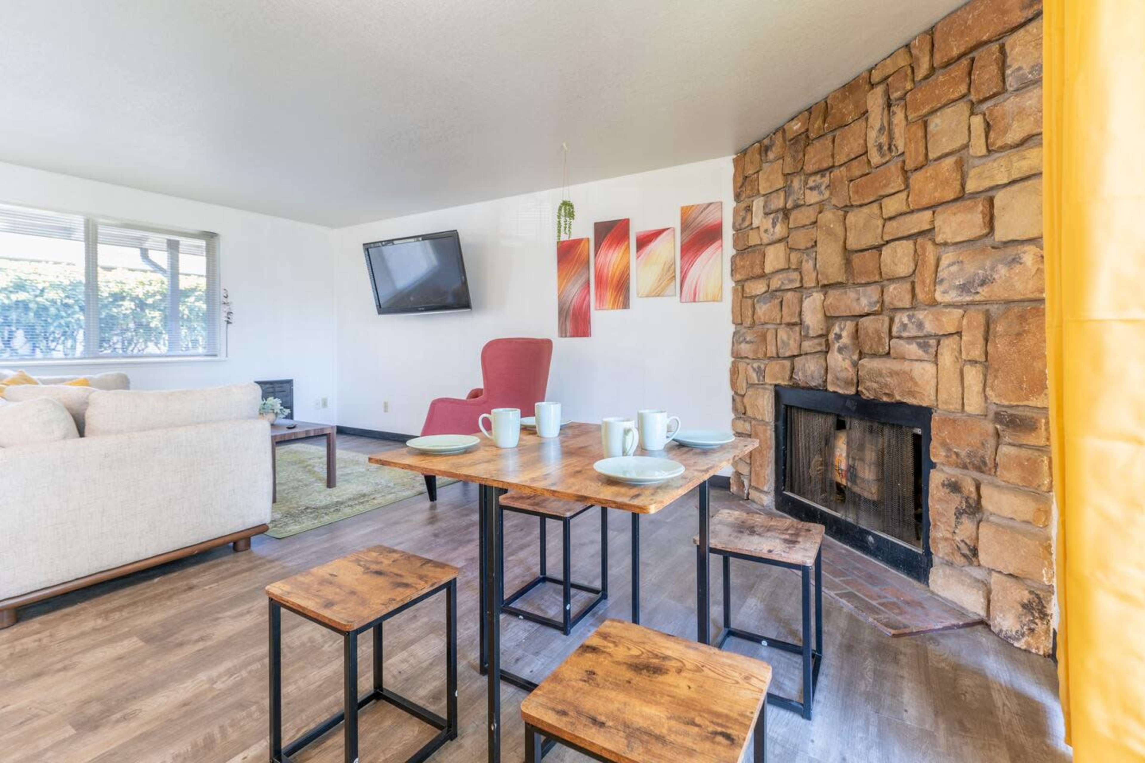 A cozy living space with a stone fireplace, a wooden dining table set for four, a television mounted on the wall, and a red armchair.