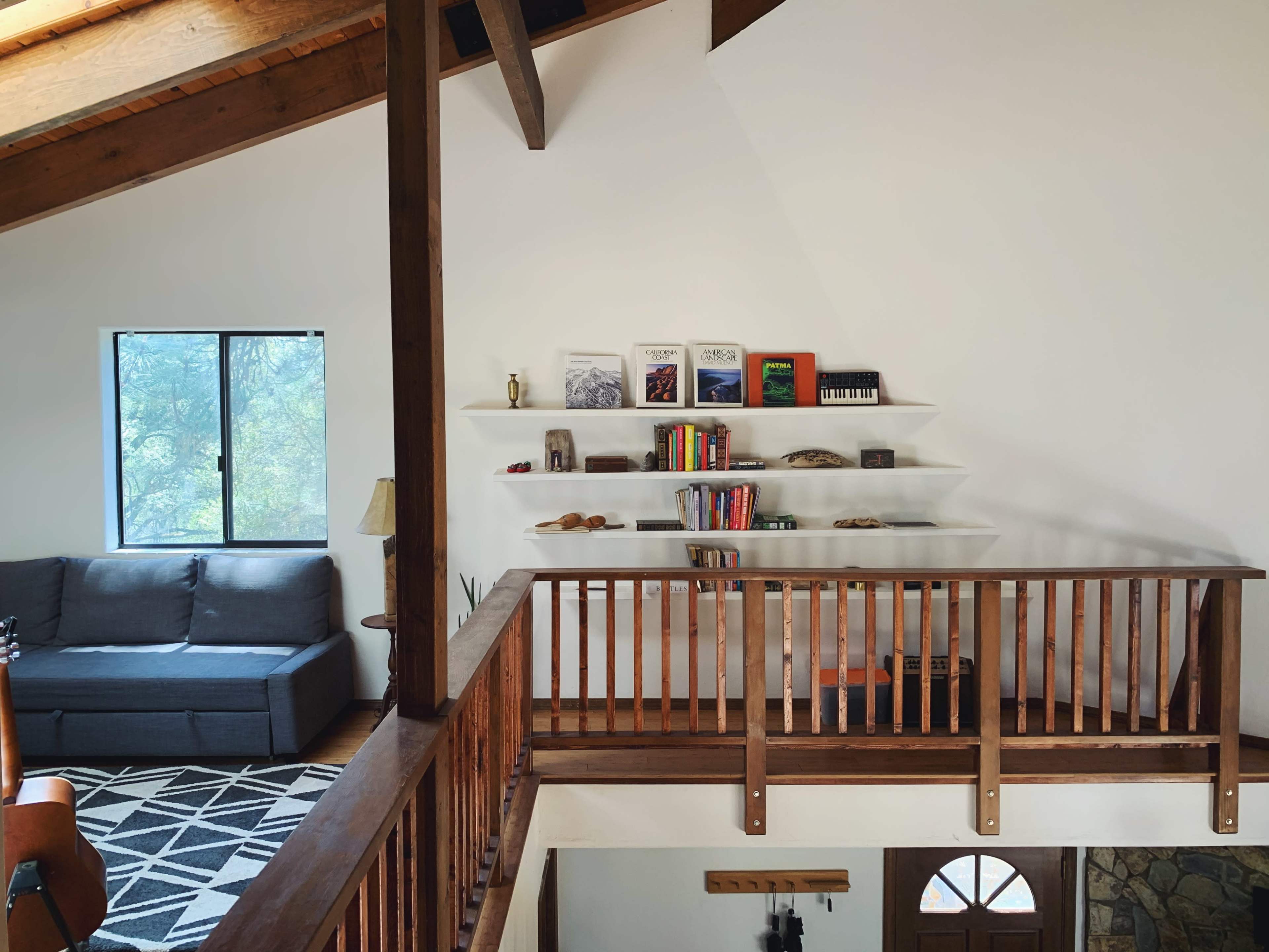 A room with a wooden balcony overlooking a living space that includes a gray couch, a bookshelf, and a door with a circular window.