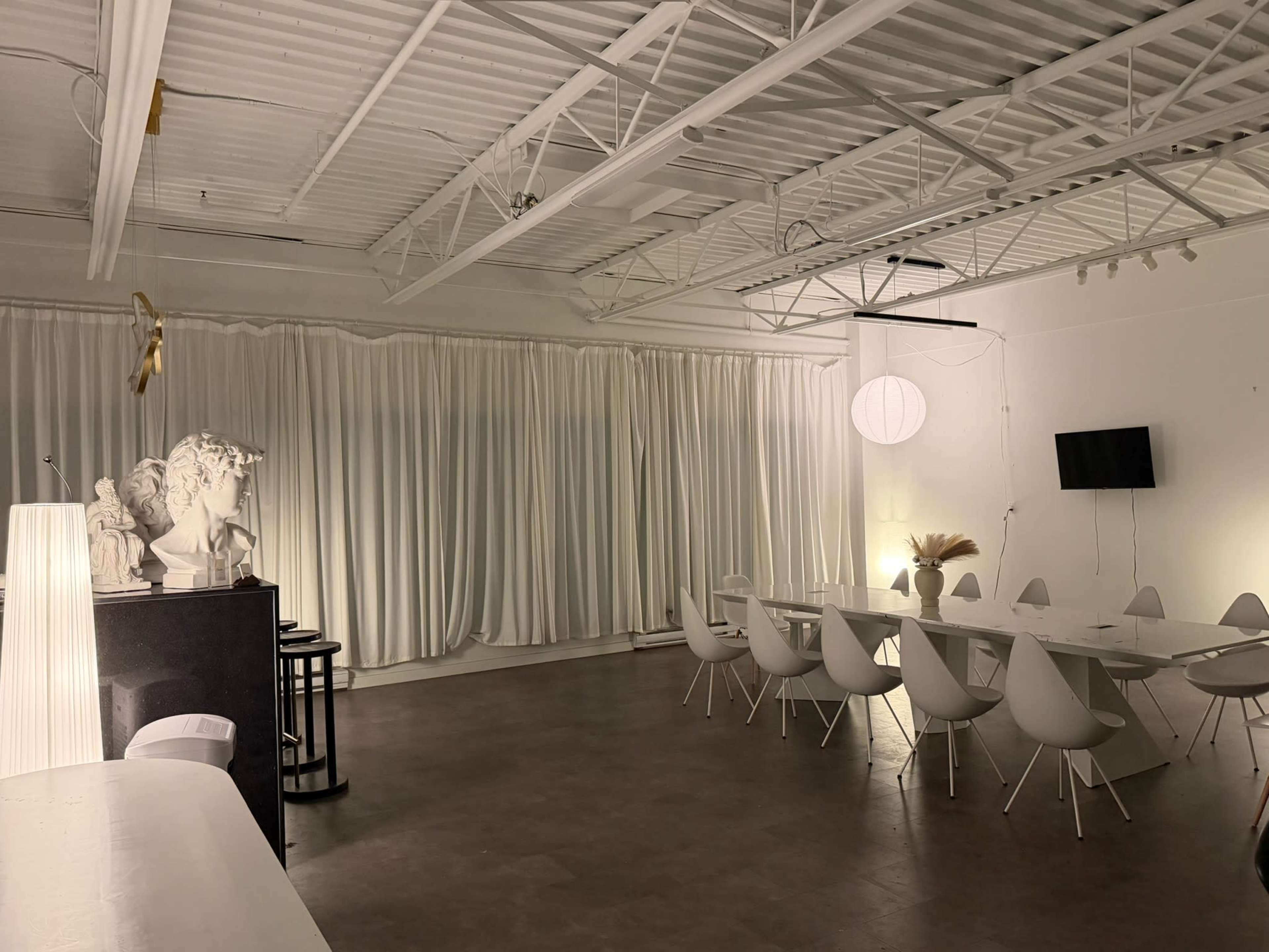 The image shows a spacious, well-lit room featuring a large white table surrounded by modern chairs, with sculptures and soft lighting among white curtains.
