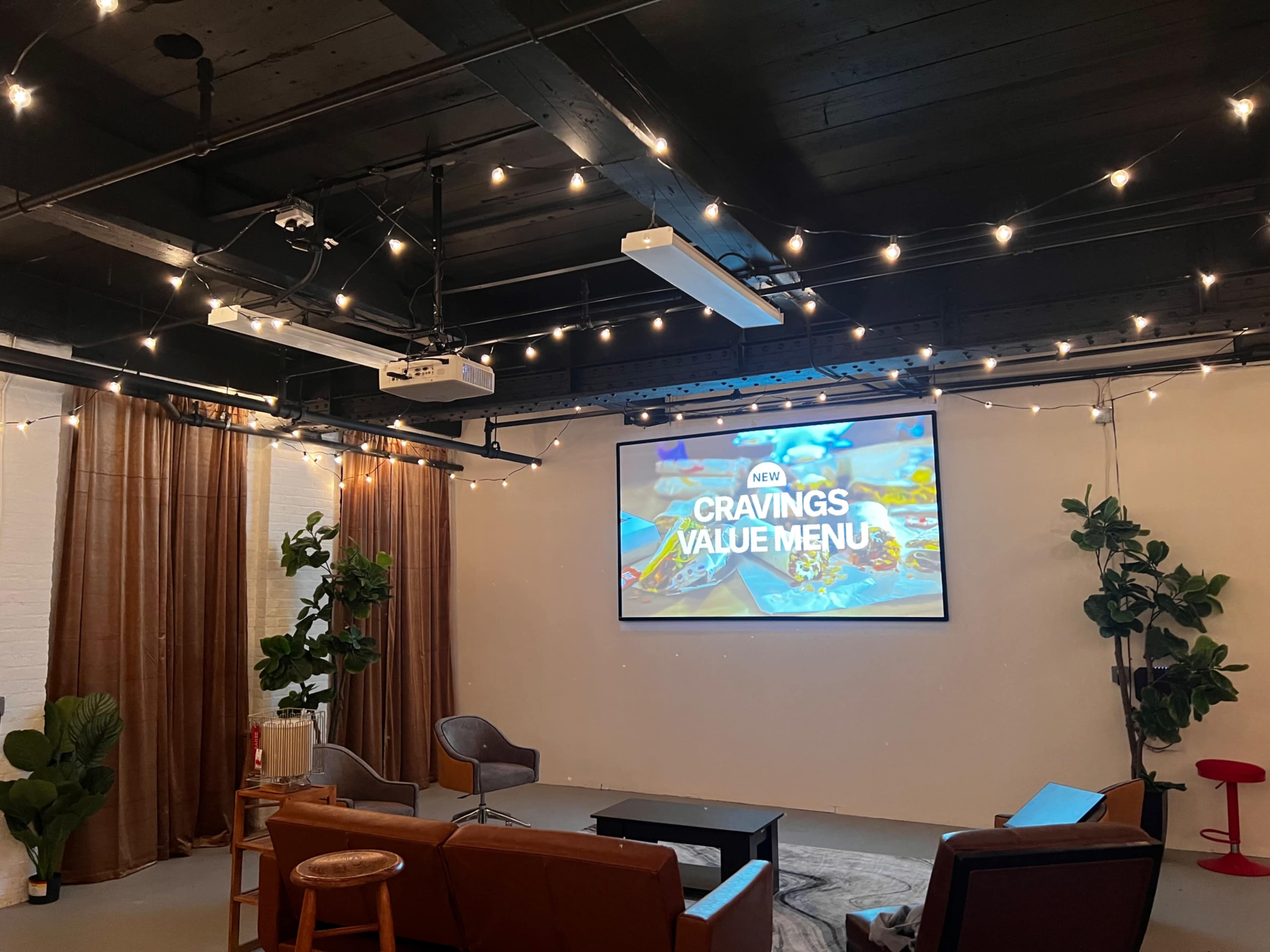 A cozy indoor space with a large projector screen displaying "NEW CRAVINGS VALUE MENU," surrounded by plants, string lights, and a few chairs and couches.