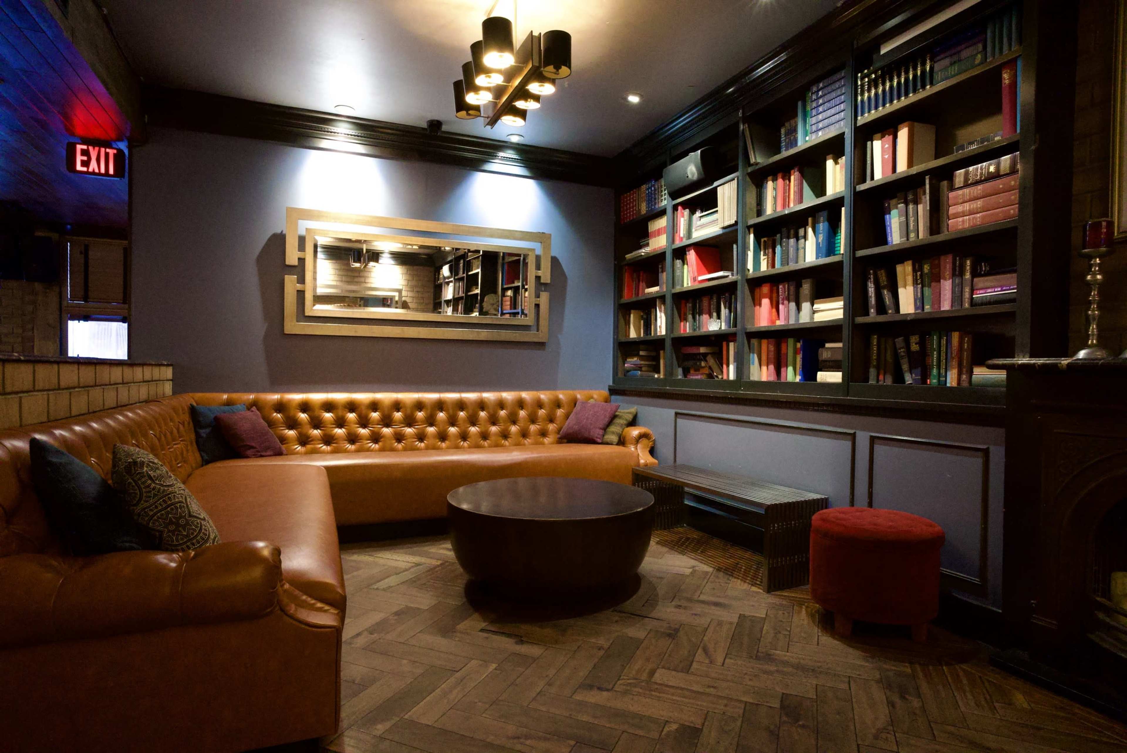 The image shows a cozy lounge area with a brown leather sofa, a round wooden coffee table, and a bookshelf filled with books, highlighted by soft lighting and a mirror on the wall.