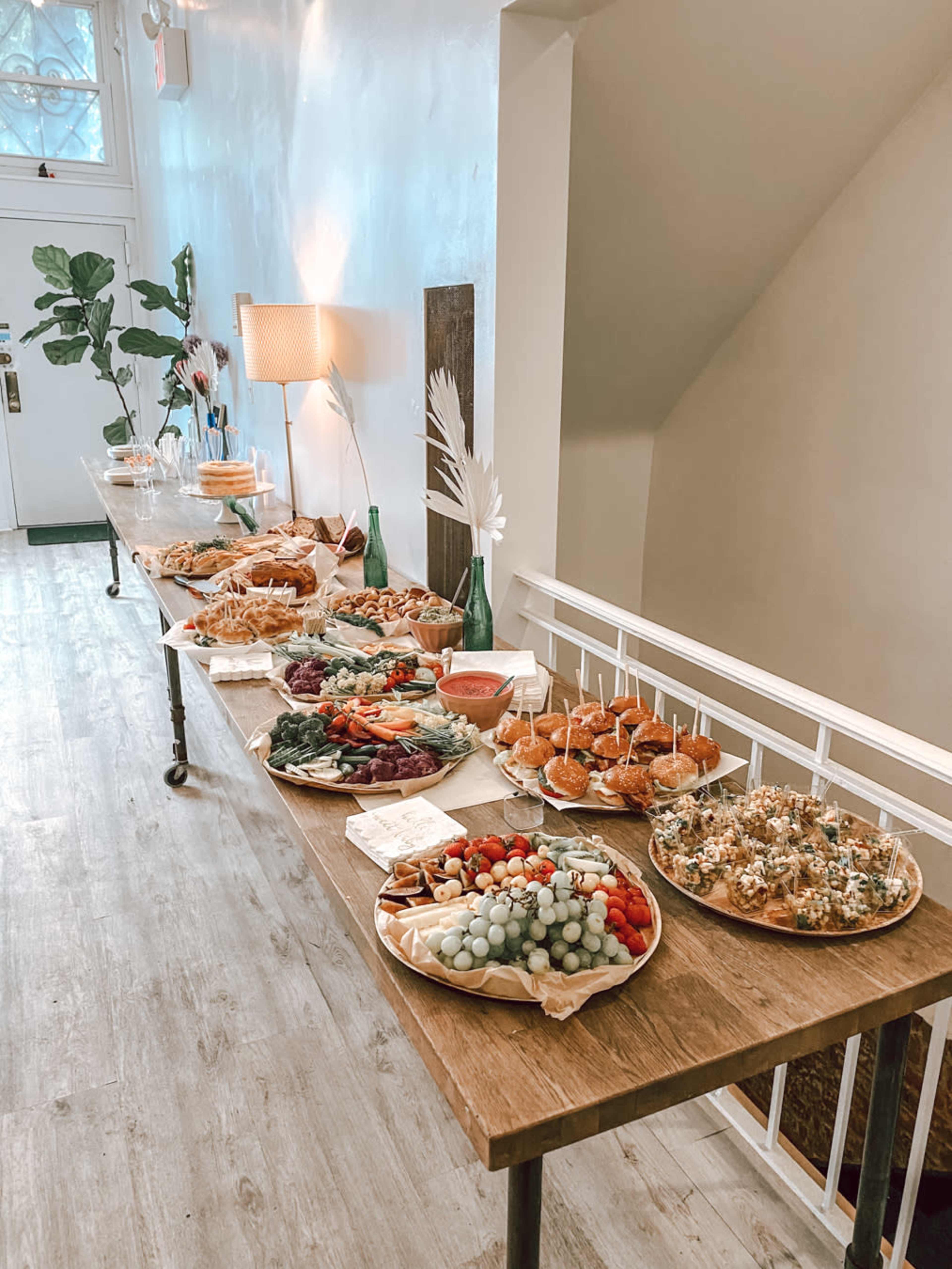 A buffet table is set with various platters of food, including fruits, vegetables, sandwiches, and pastries, alongside decorative elements.
