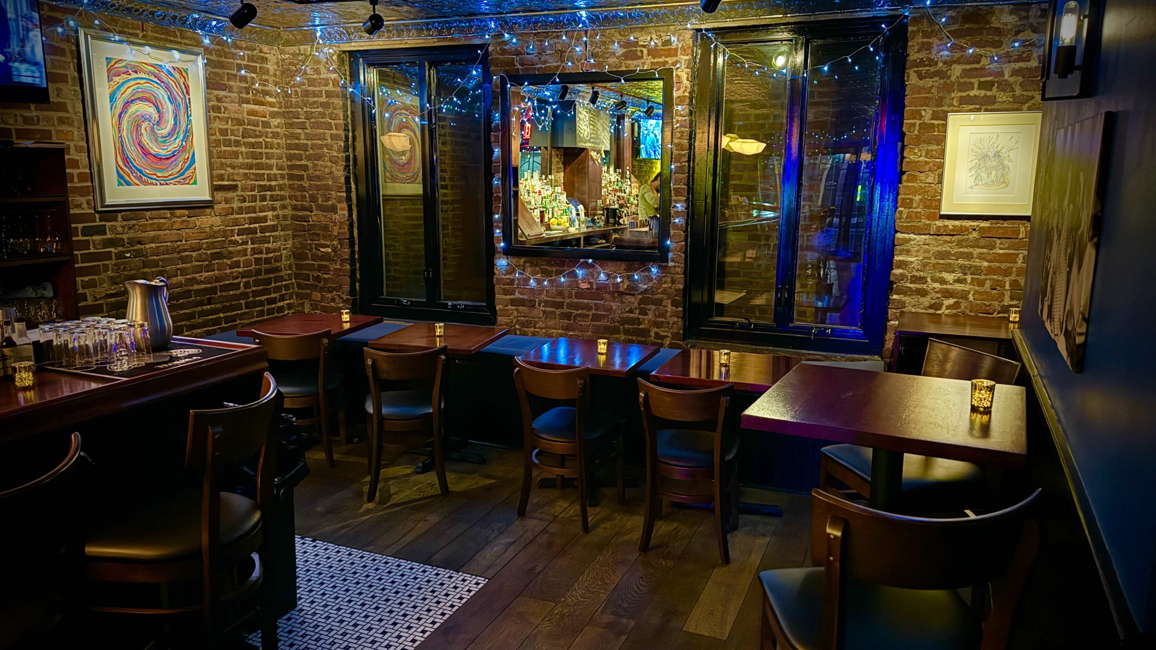 A dimly lit bar interior with brick walls, wooden tables and chairs, and colorful lights strung across the windows.