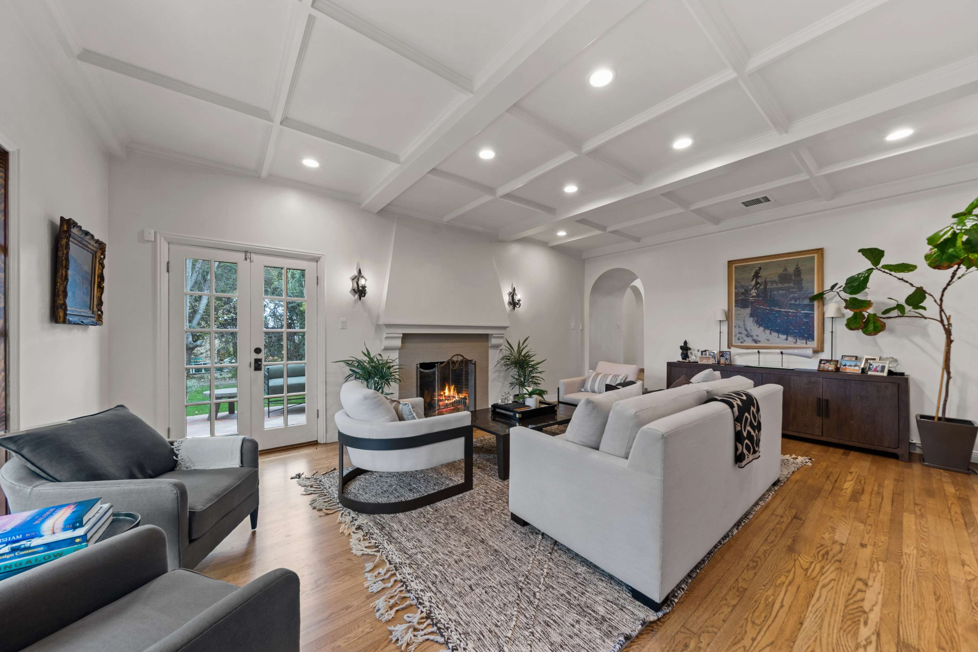 The image shows a modern living room with a coffered ceiling, a fireplace, two sofas, and large windows leading to an outdoor view.