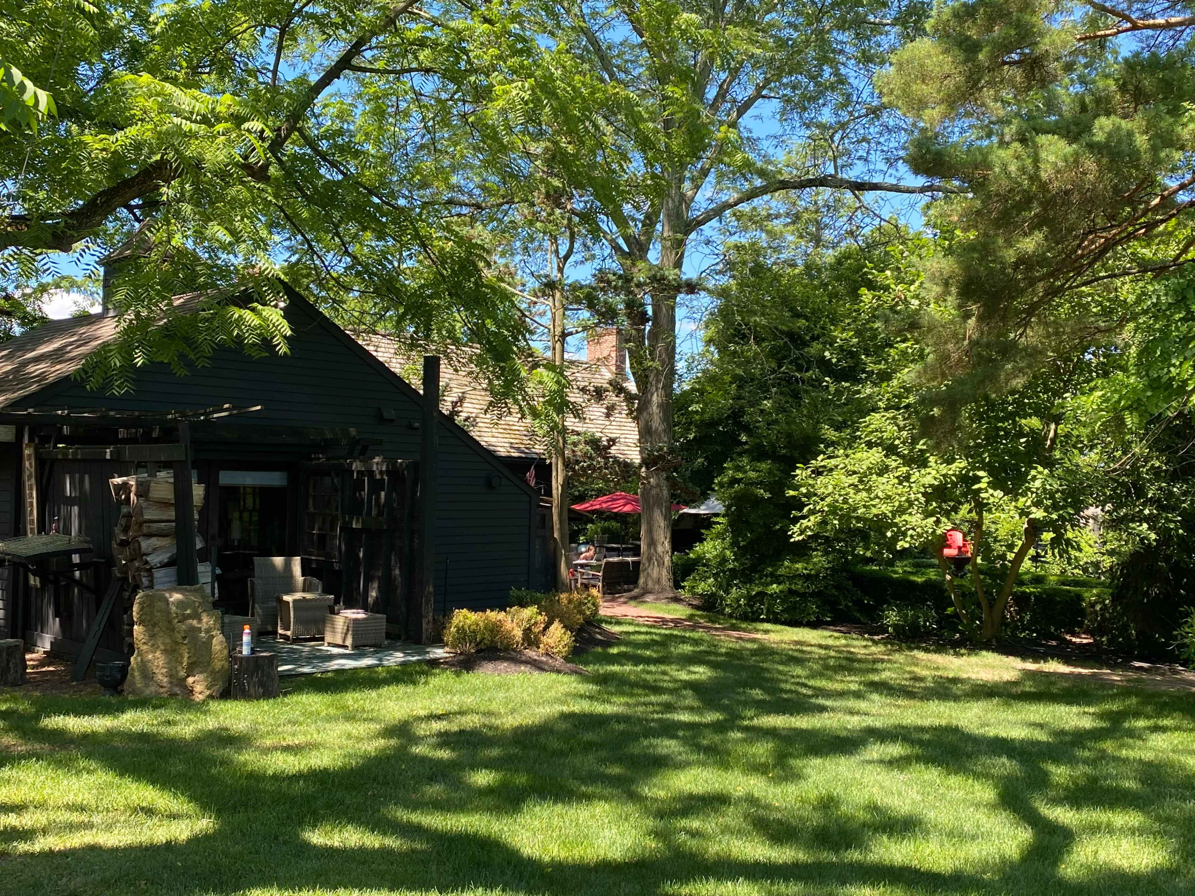 A rustic black wooden building surrounded by lush green grass and trees, with a hint of red from an outdoor umbrella visible in the background.