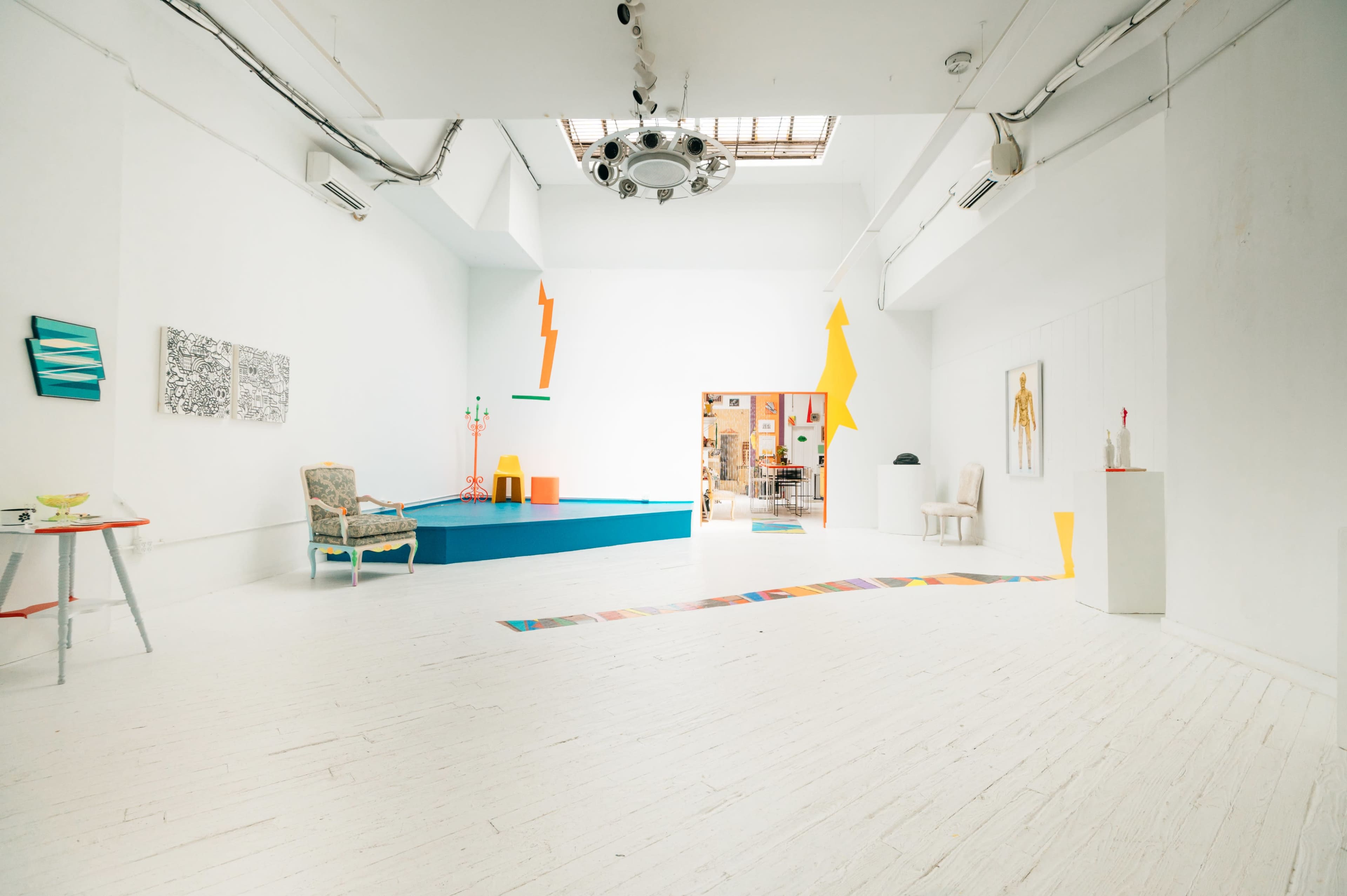 A bright, spacious gallery with white walls, colorful geometric decorations, and a few pieces of furniture arranged around a vibrant blue platform.