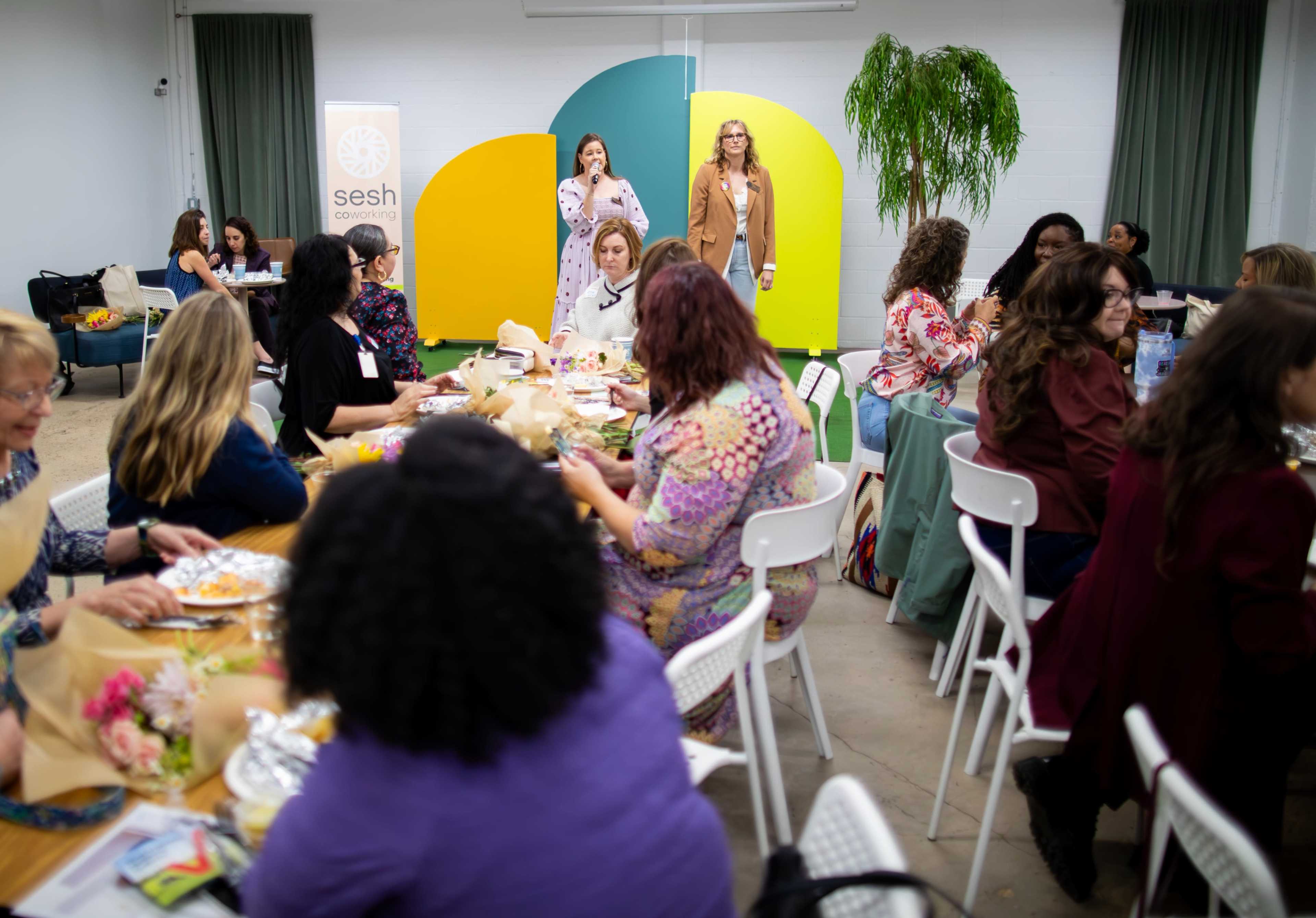 A group of women are gathered around tables enjoying food and conversation, while two speakers address the audience in a brightly decorated room.
