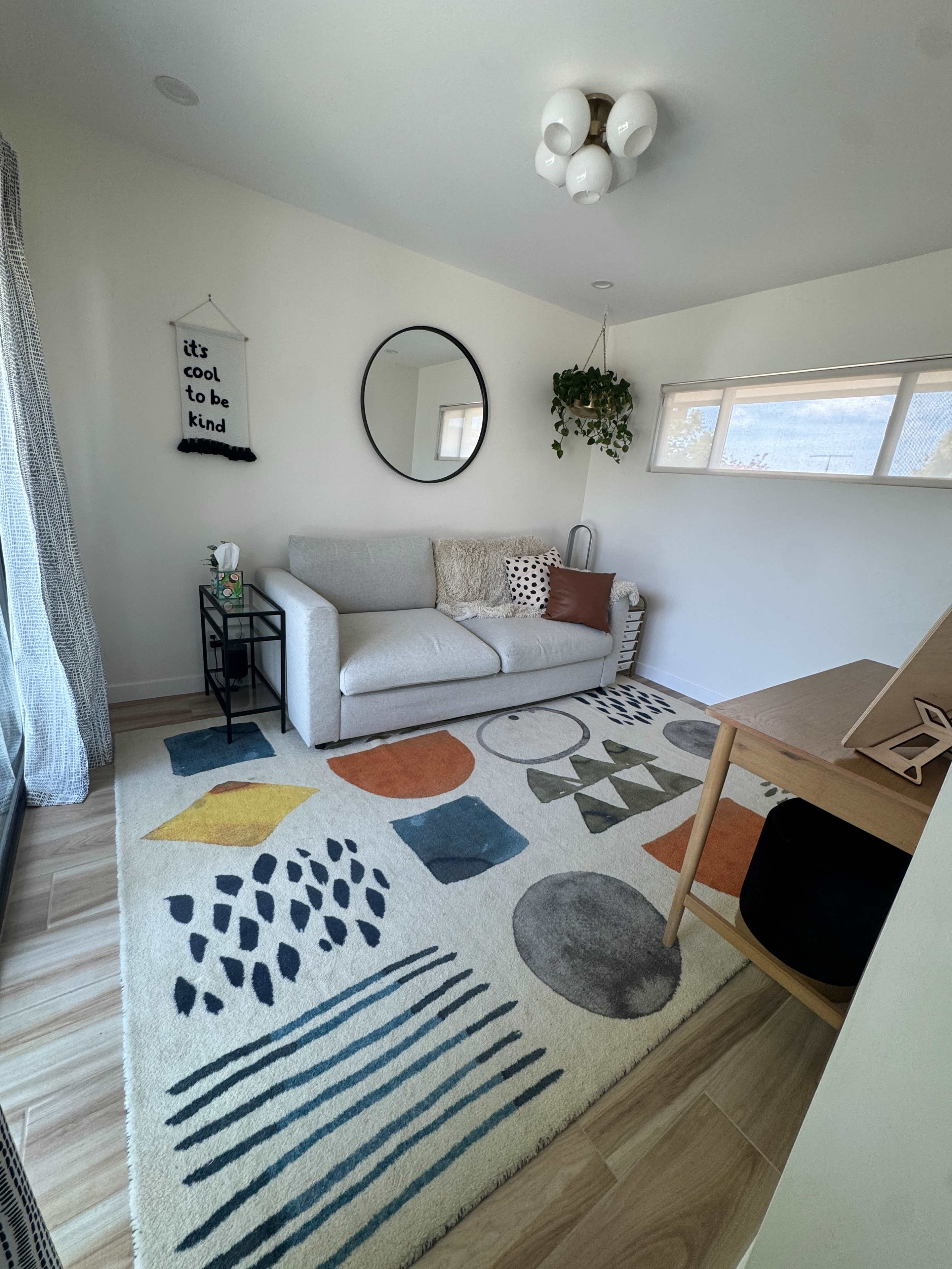 The image shows a bright living room featuring a gray sofa, a round mirror, and a patterned area rug with bold geometric shapes, accompanied by a small wooden desk and a hanging plant.
