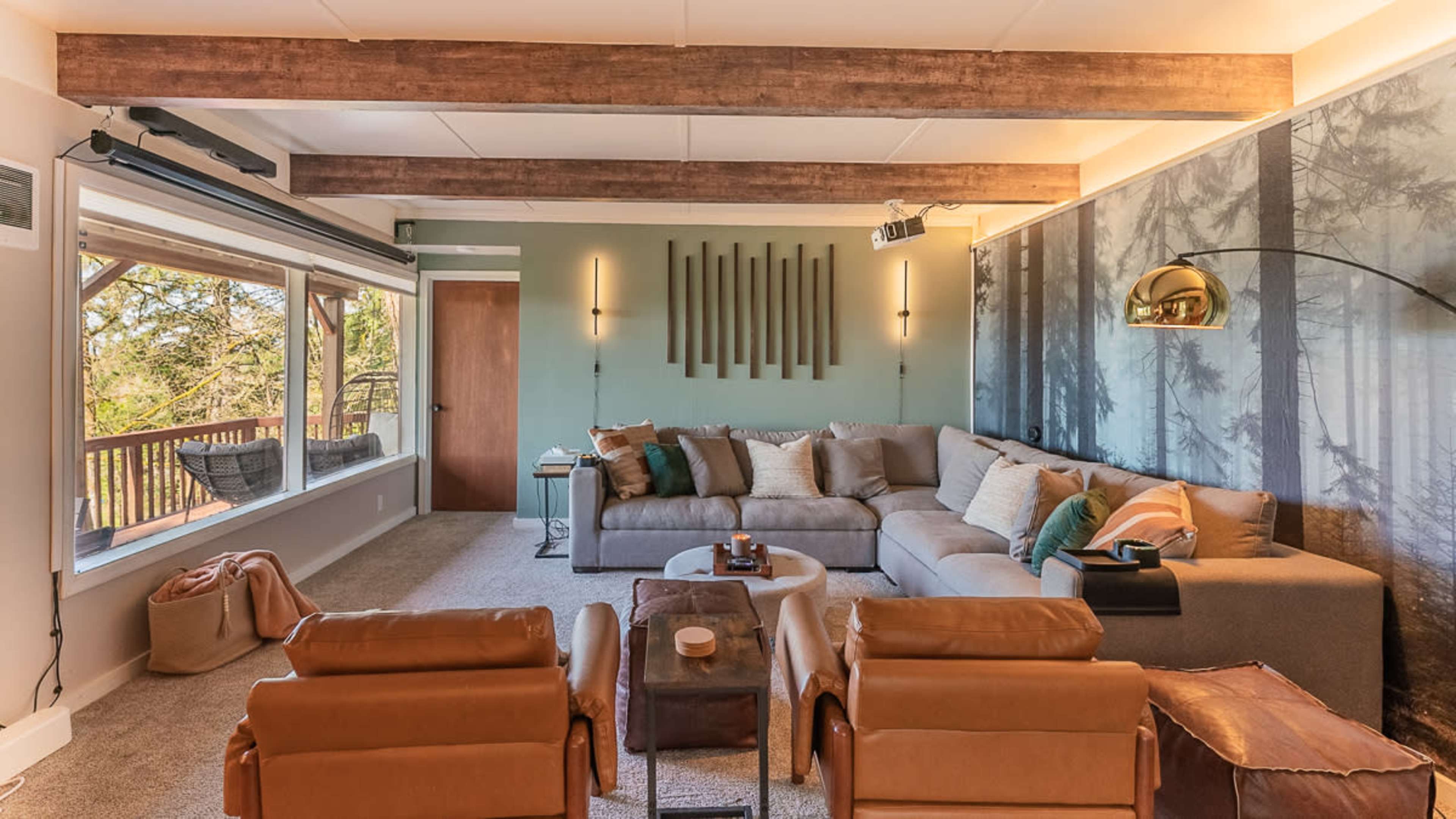 A modern living room features a large sectional sofa, two leather chairs, and a window that opens to a forest view.