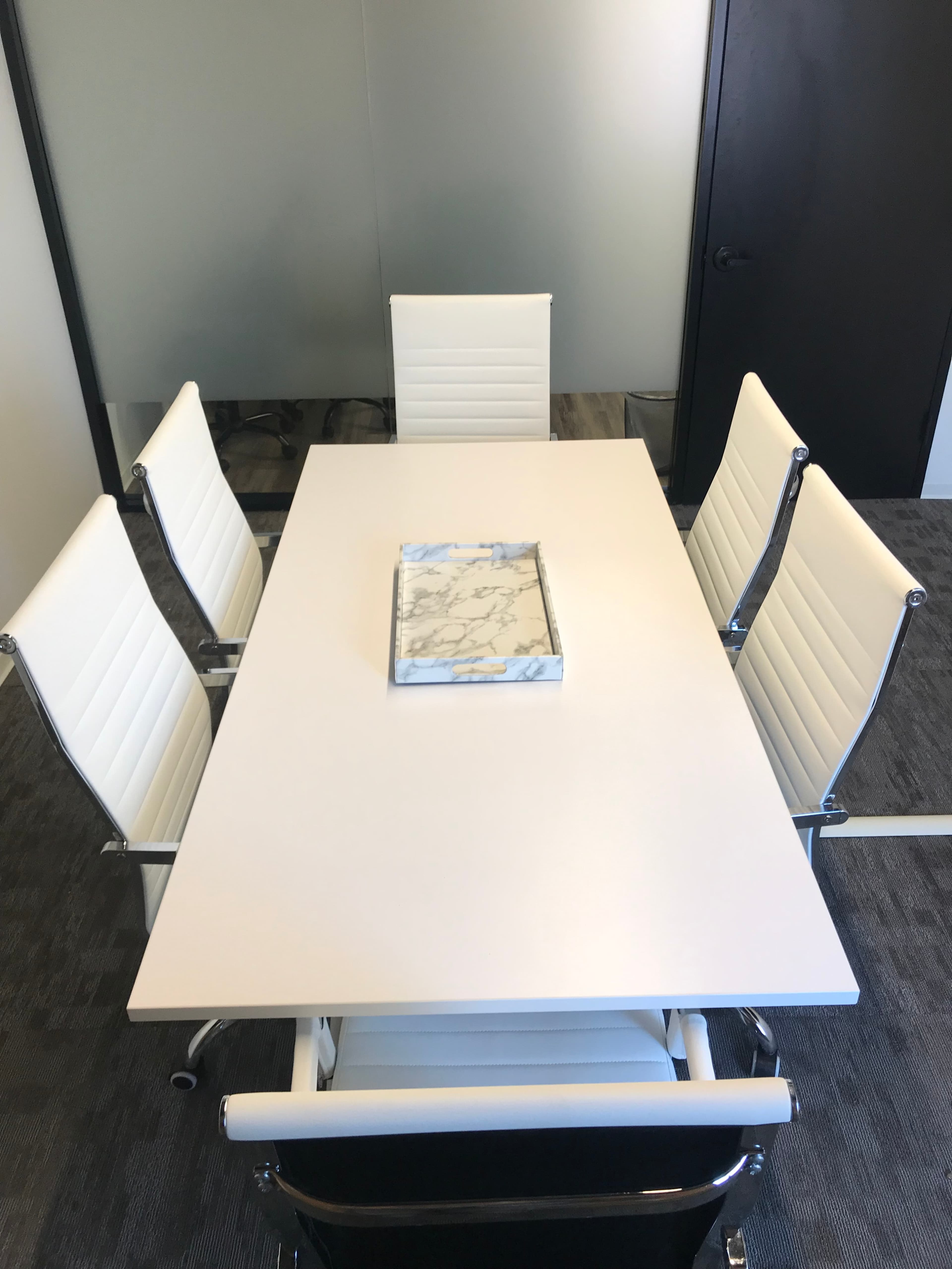 A modern conference room features a white rectangular table surrounded by four white chairs, with a marble-patterned tray positioned in the center.