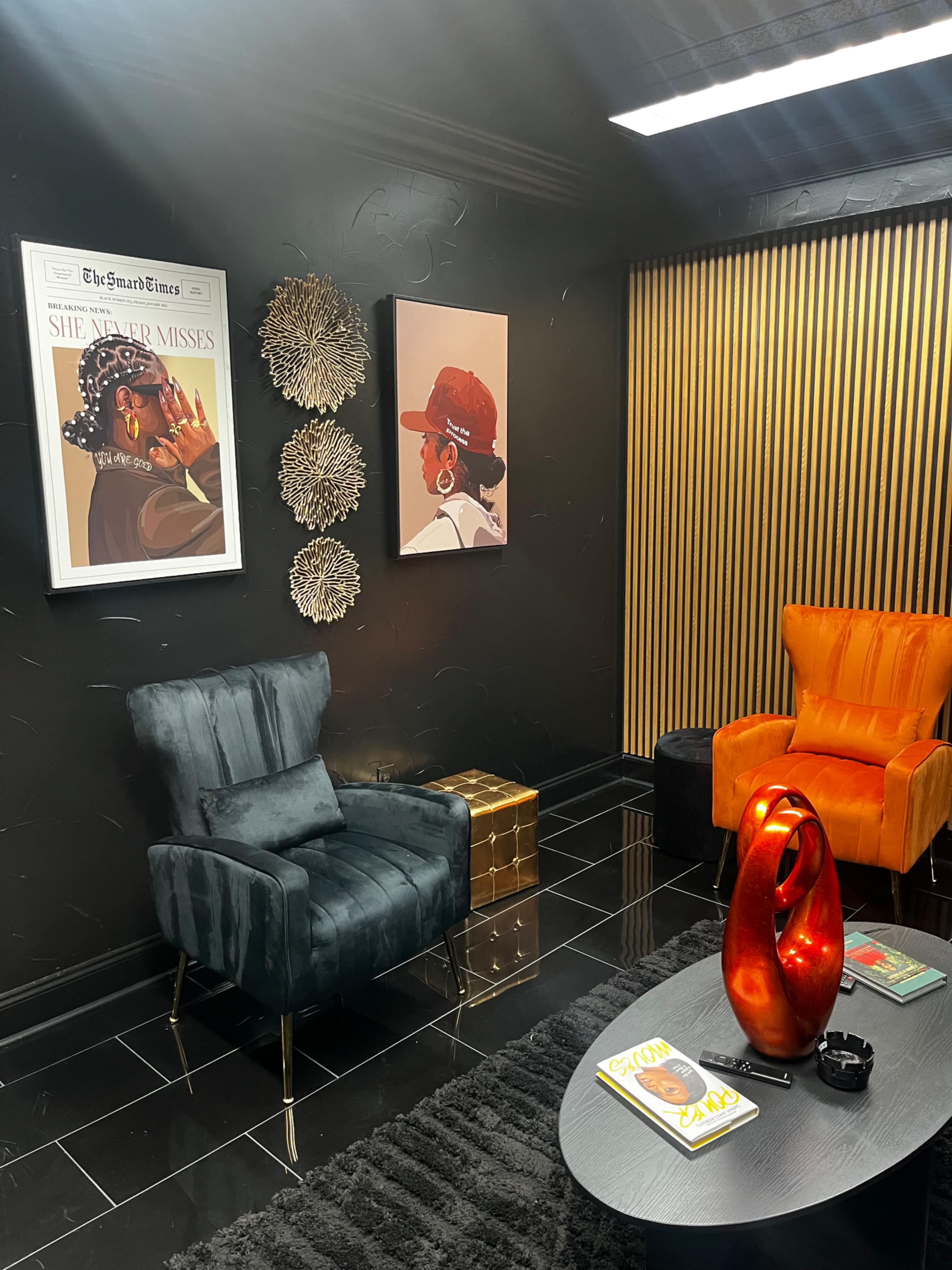 The image shows a modern sitting area with two armchairs, one orange and one teal, along with framed artwork and decorative wall elements on black walls.