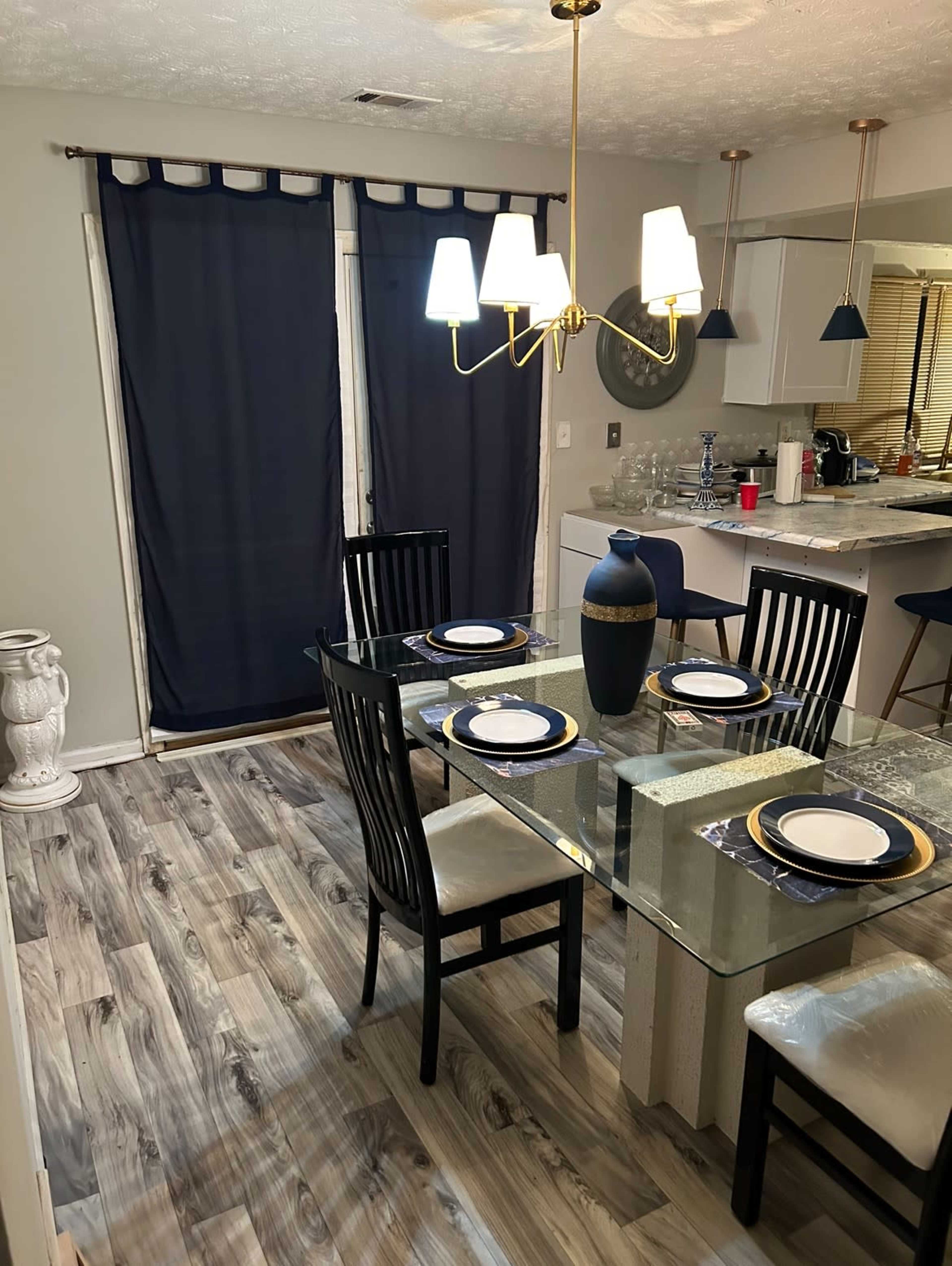 A dining area features a glass table with four black chairs, a vase centerpiece, and a backdrop of navy curtains.
