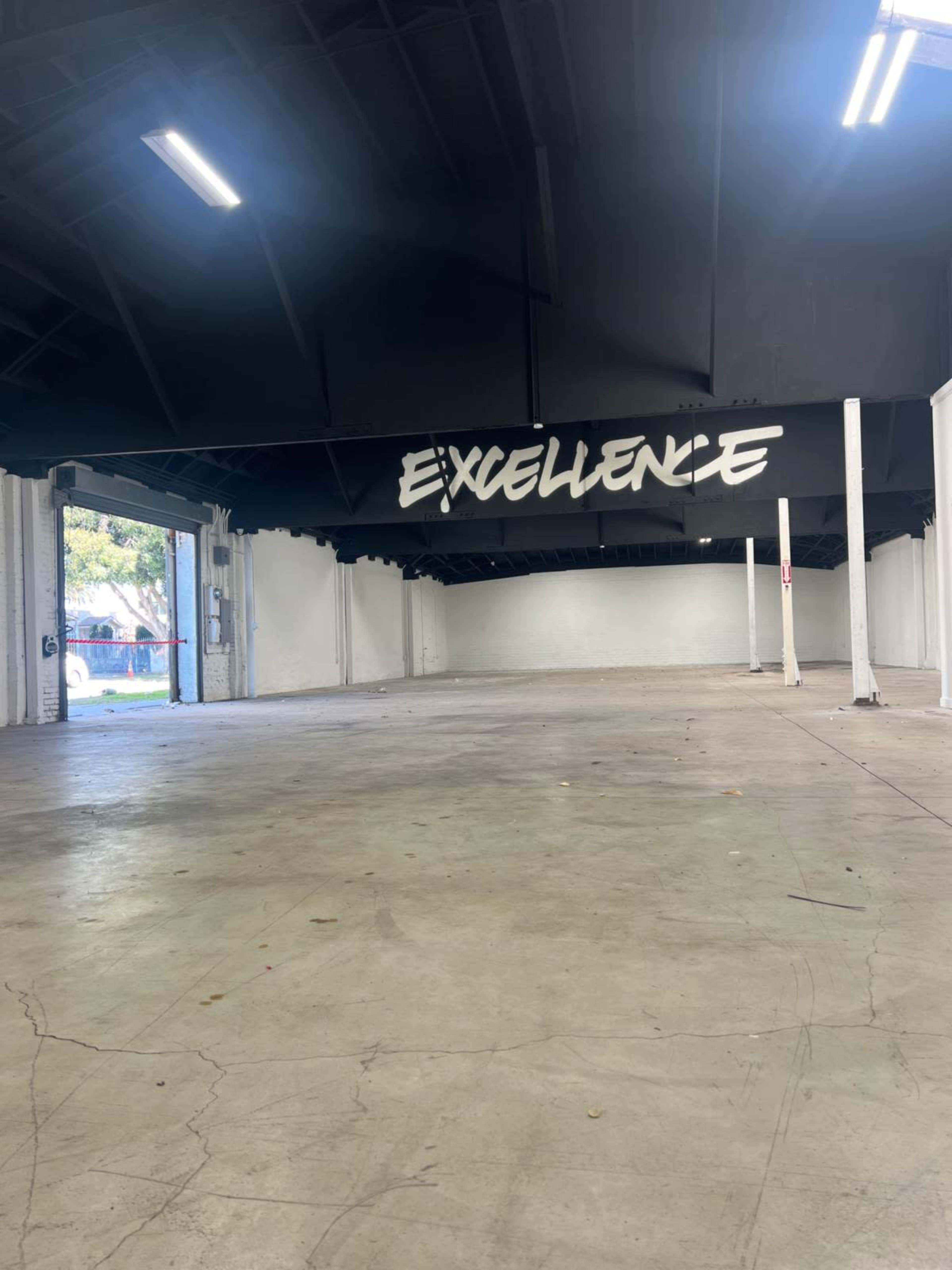 The image shows an empty industrial space with a large sign that reads "EXCELLENCE" overhead on a black ceiling.