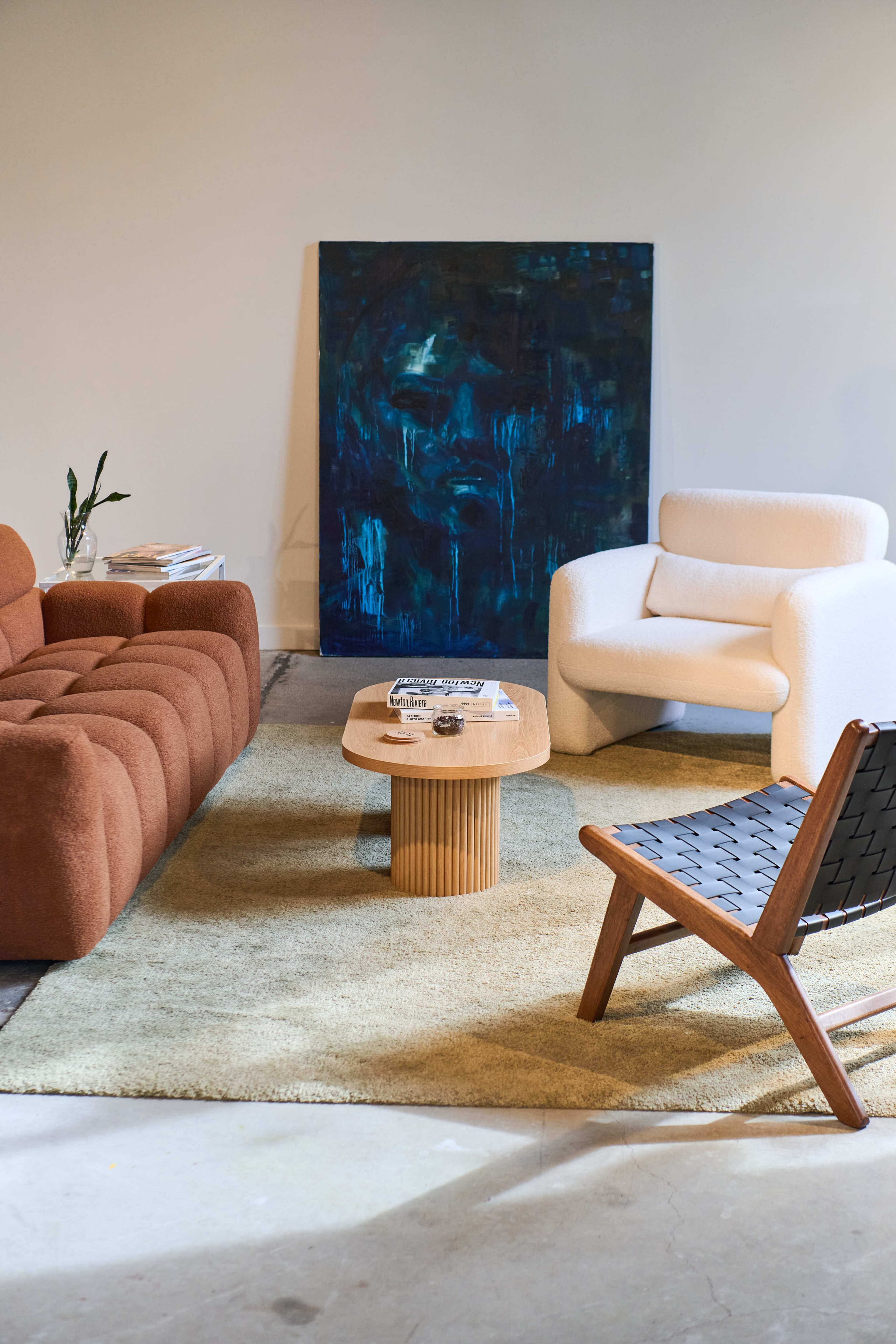The image shows a modern living room with a brown sofa, a white armchair, a round wooden coffee table, and a textured area rug, set against a wall with an abstract blue painting.