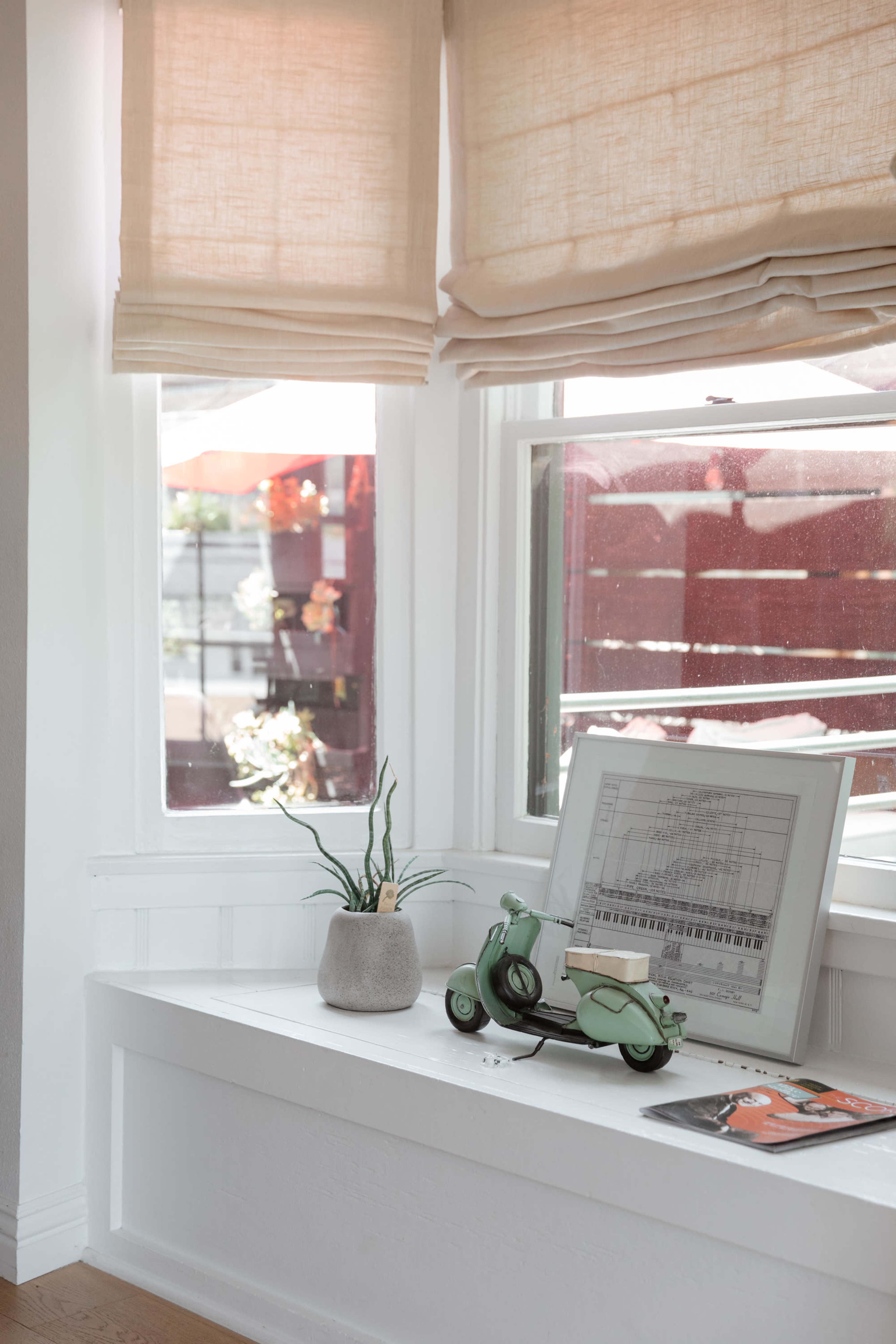 A window seat features a small potted plant, an illustration in a frame, and a vintage toy scooter, with light filtering through the fabric shades.