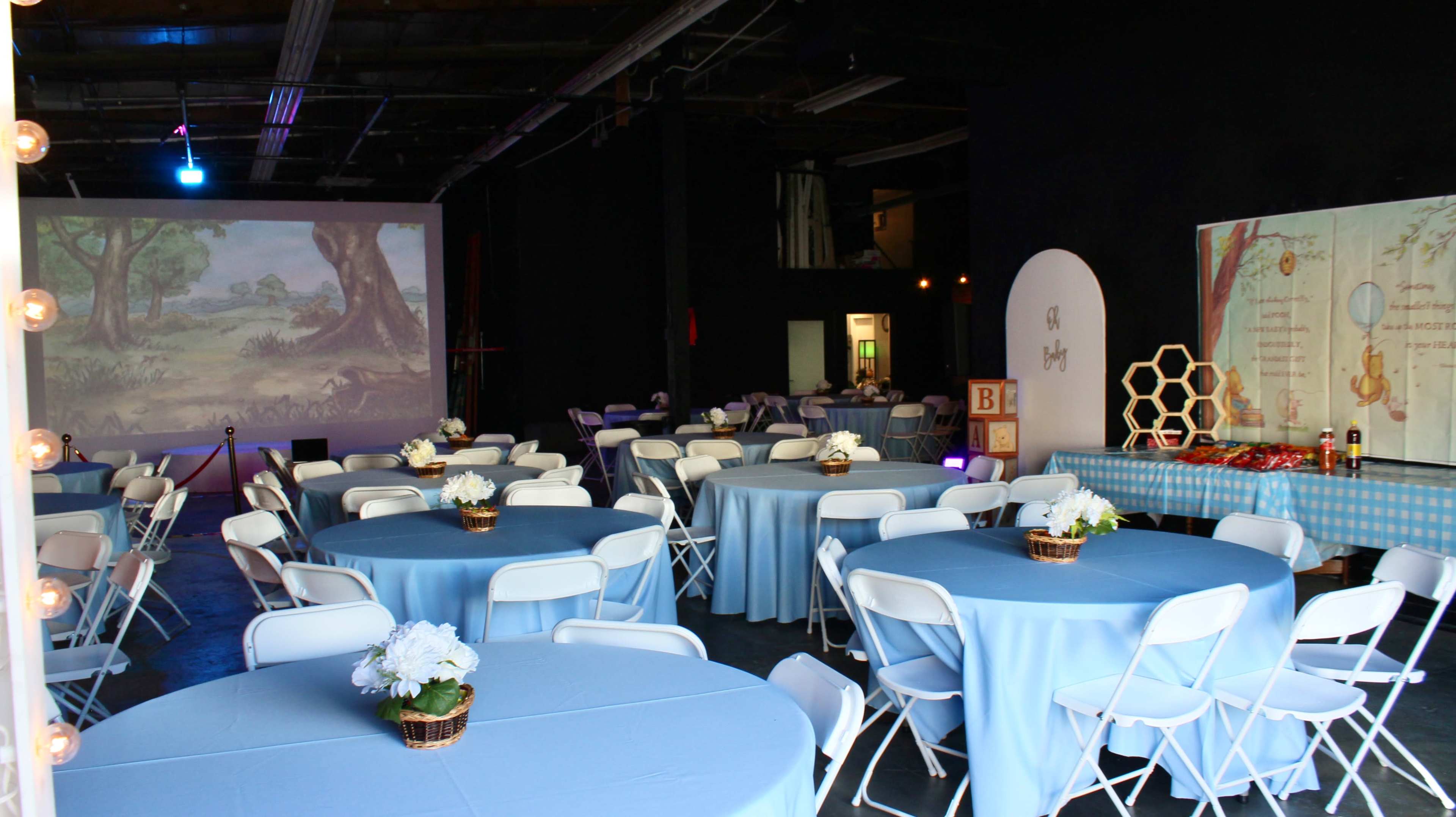 The image shows a decorated indoor event space with tables covered in light blue tablecloths, surrounded by white chairs, and a large projection screen displaying a nature scene in the background.
