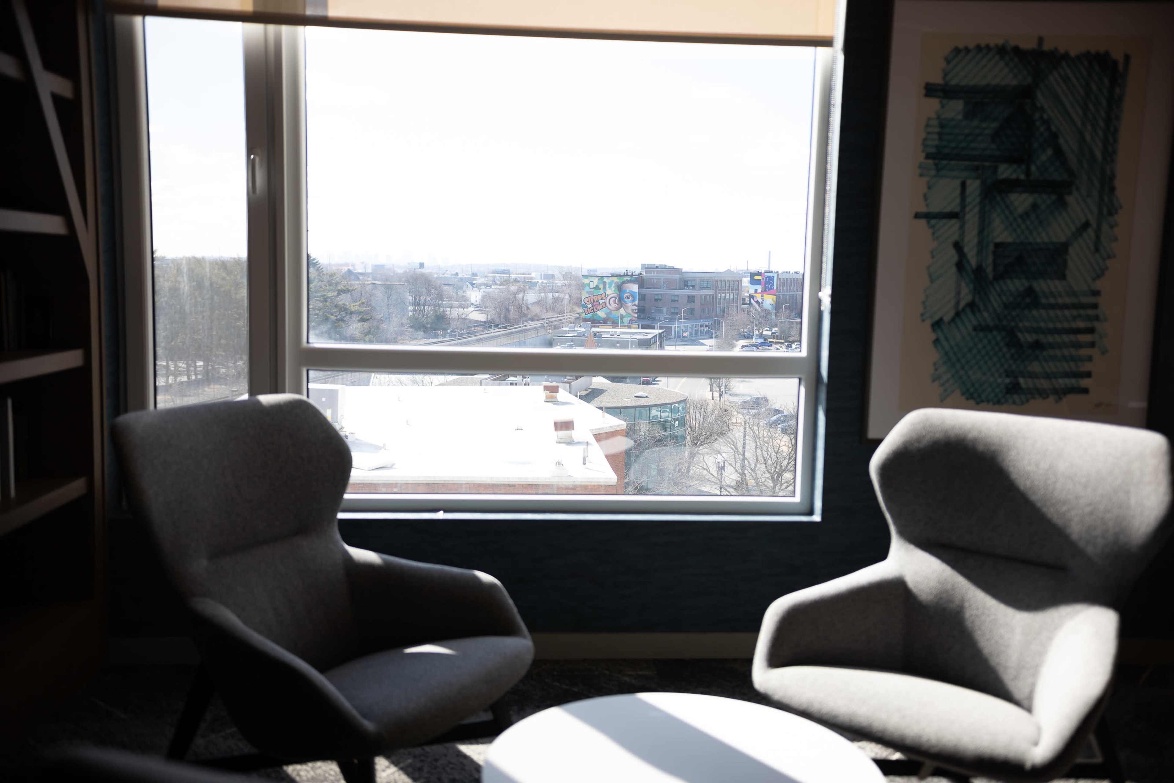 The image shows two gray armchairs positioned near a large window, overlooking an outdoor view of a cityscape with buildings and greenery.