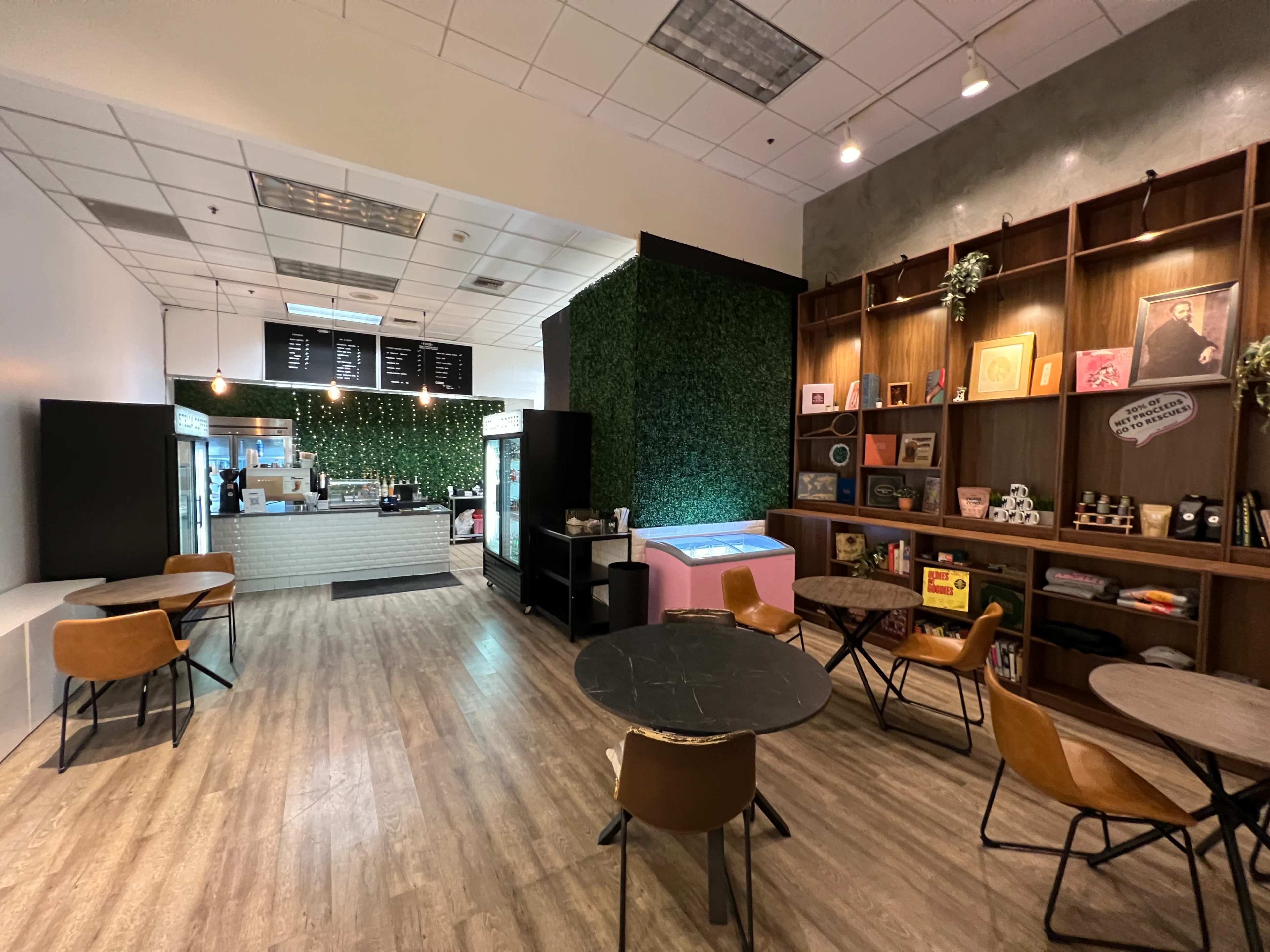 The image shows a modern café with a seating area featuring wooden tables and chairs, a green wall, and a counter displaying a menu and refrigerated items.