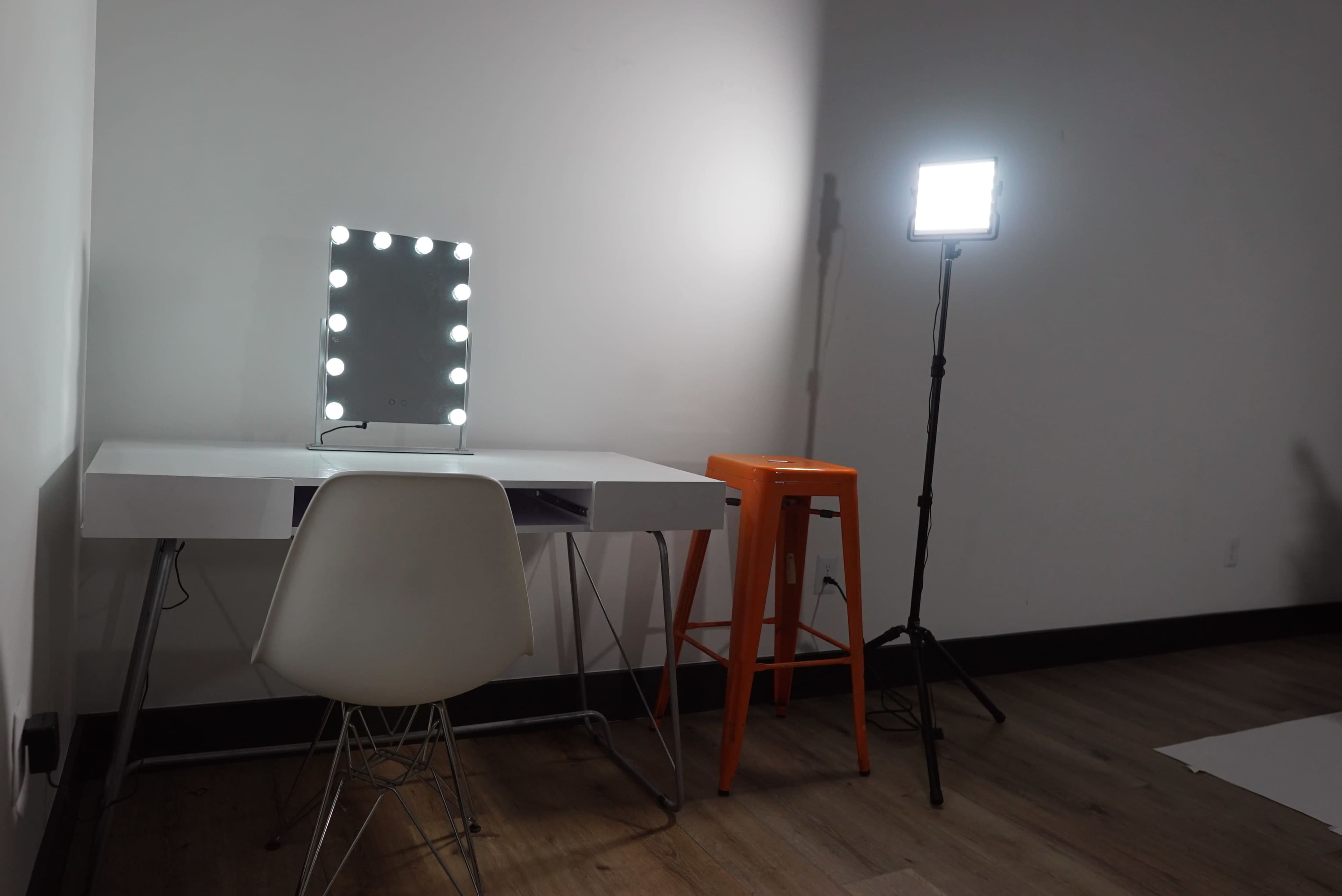 A simple makeup vanity with a mirror and lights sits next to an orange bar stool and a standing light in a well-lit room.