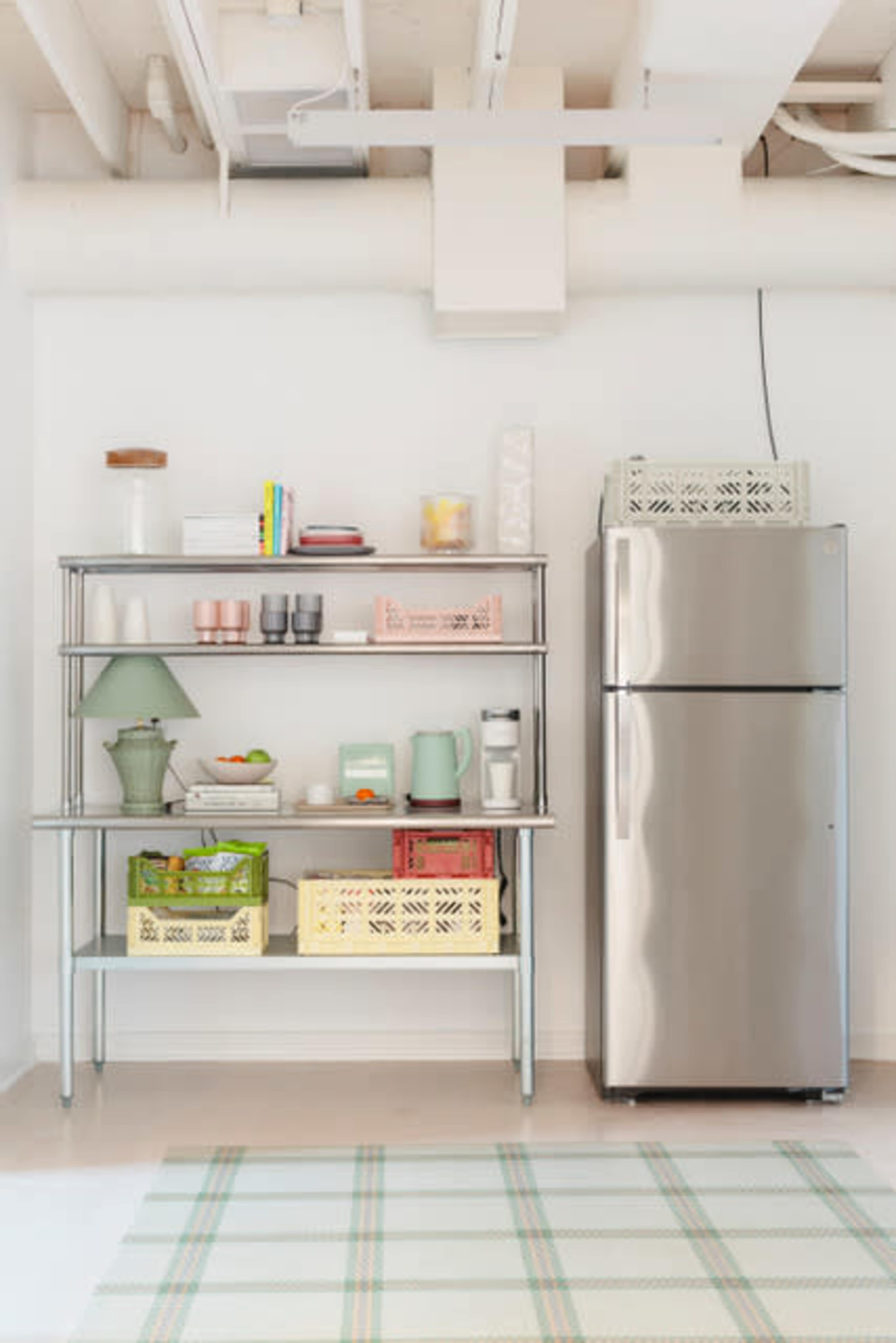 A metal shelving unit with various kitchen items sits next to a stainless steel refrigerator in a bright, open space.