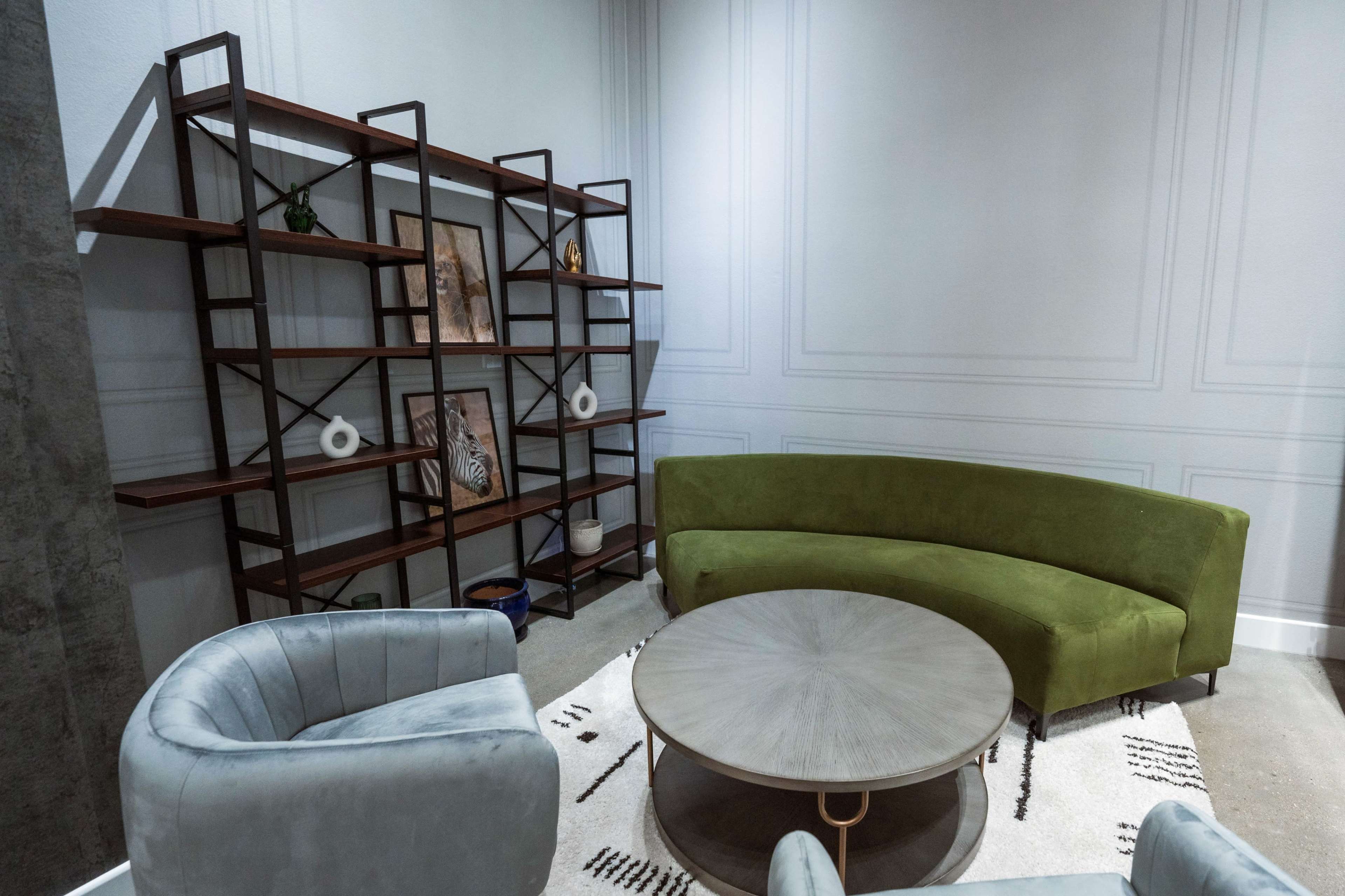 The image shows a modern seating area with a green curved sofa, two light gray armchairs, and a circular wooden coffee table, backed by a geometric wooden shelf displaying decorative items.