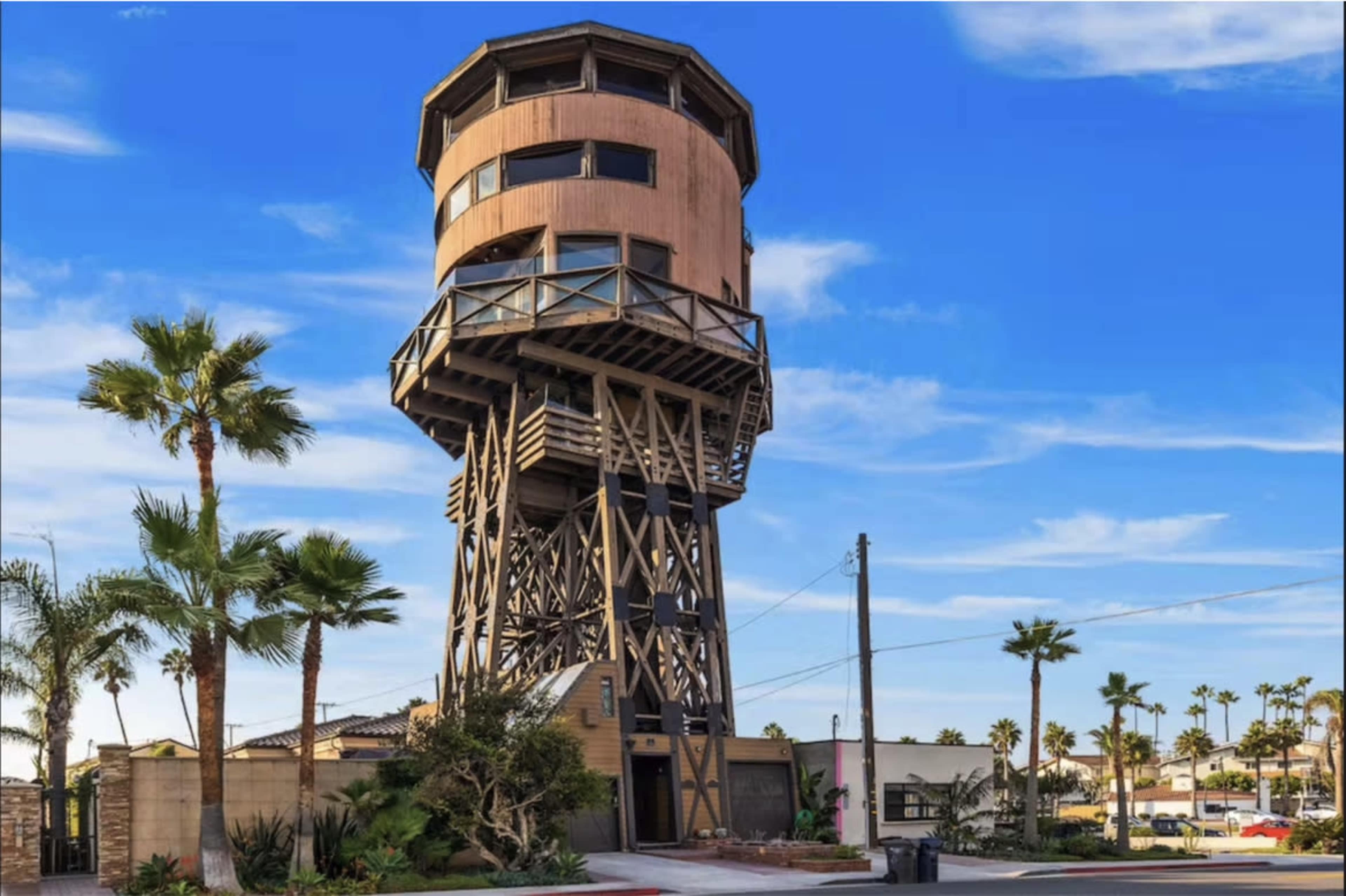 A tall wooden structure resembling a water tower, standing on a support framework with palm trees in the foreground against a blue sky.