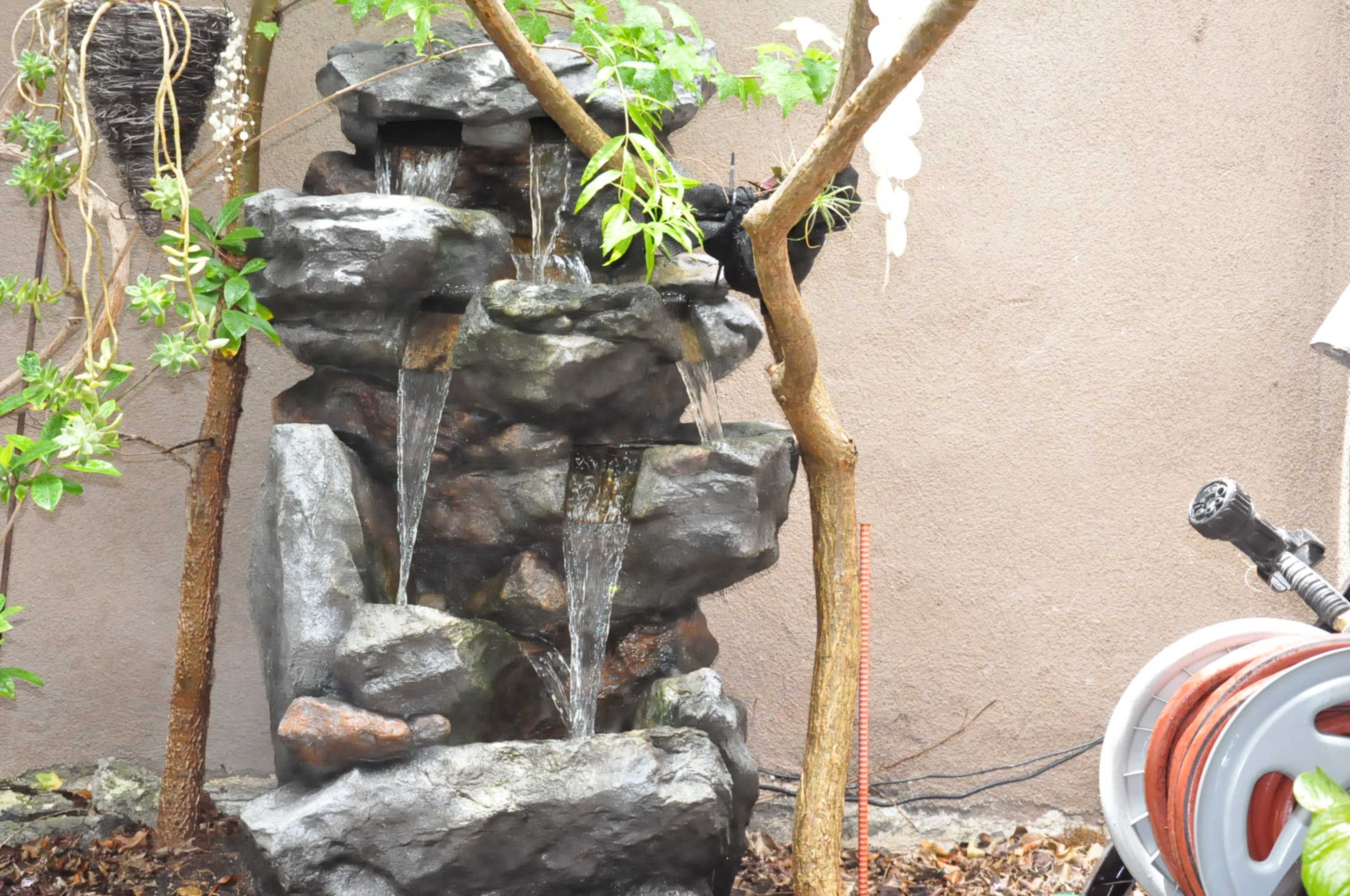 A stone water fountain with multiple tiers flows gently in a garden area beside a garden hose.