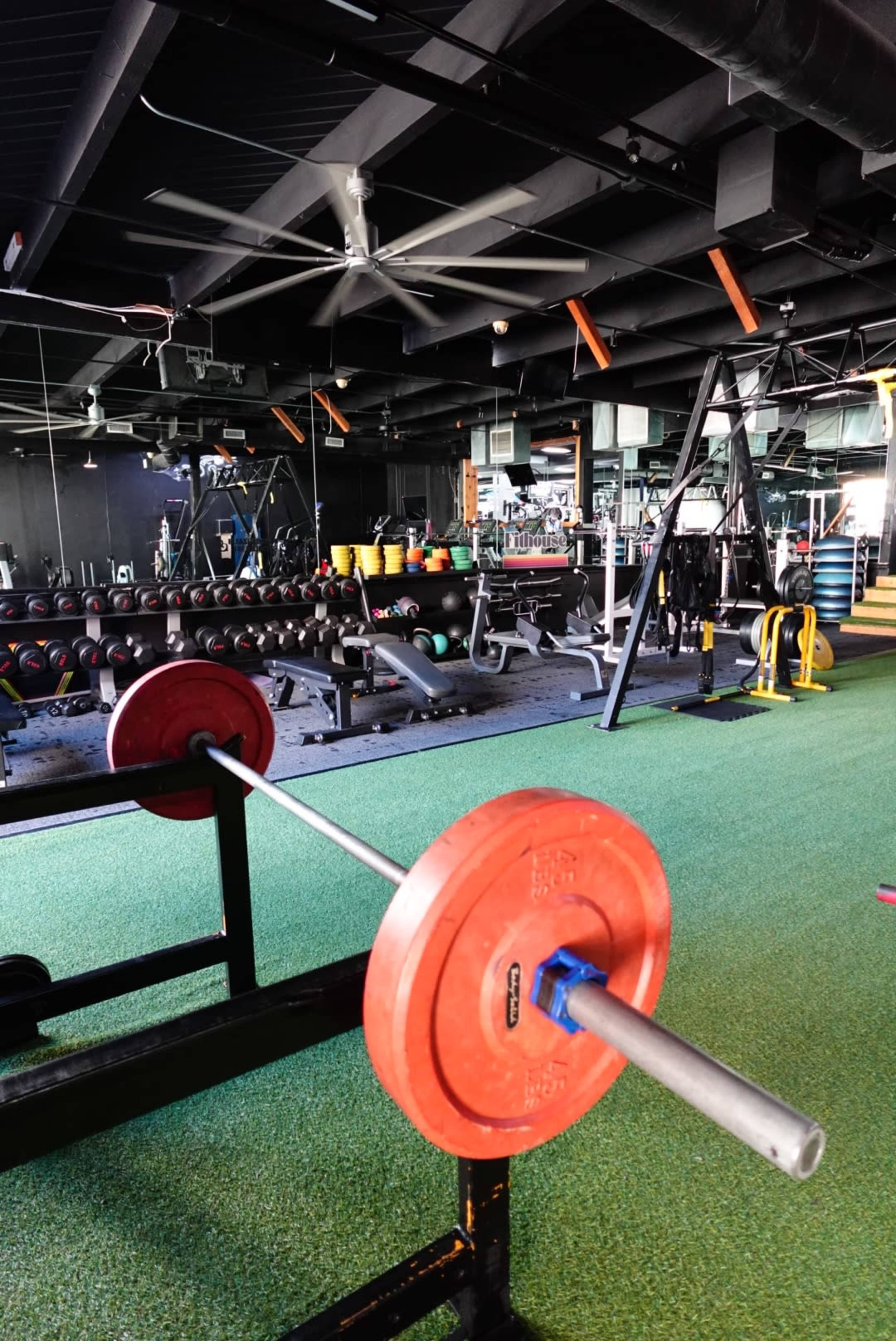 The image shows a gym interior with weightlifting equipment, including a barbell and various weights, arranged in a spacious area with artificial turf flooring.