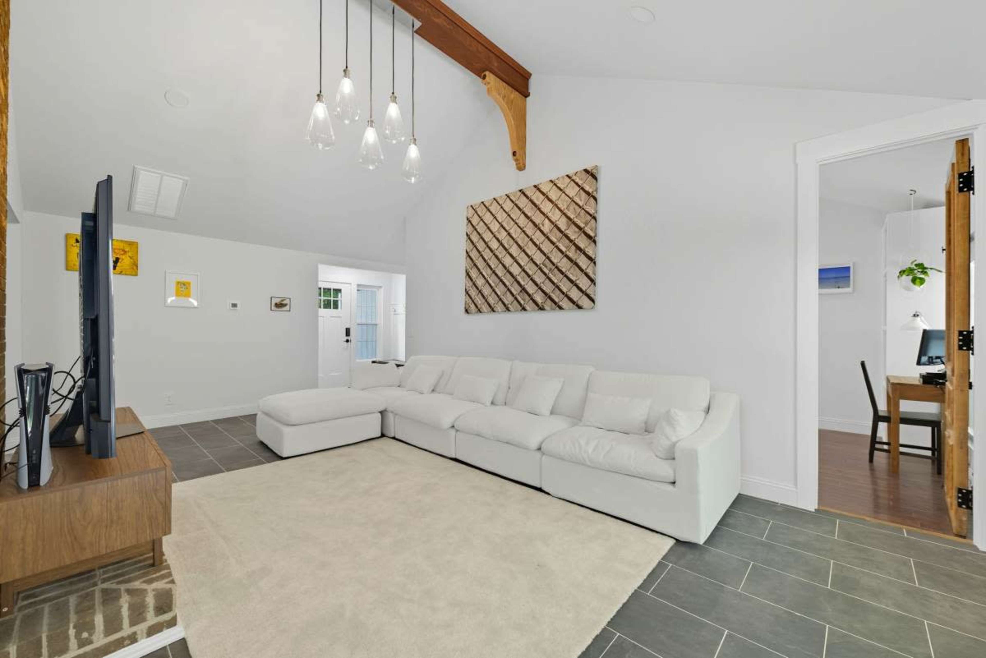 The image shows a modern living room with a white sectional sofa, a flat-screen TV on a wooden console, and a light-colored area rug under a ceiling with wooden beams.