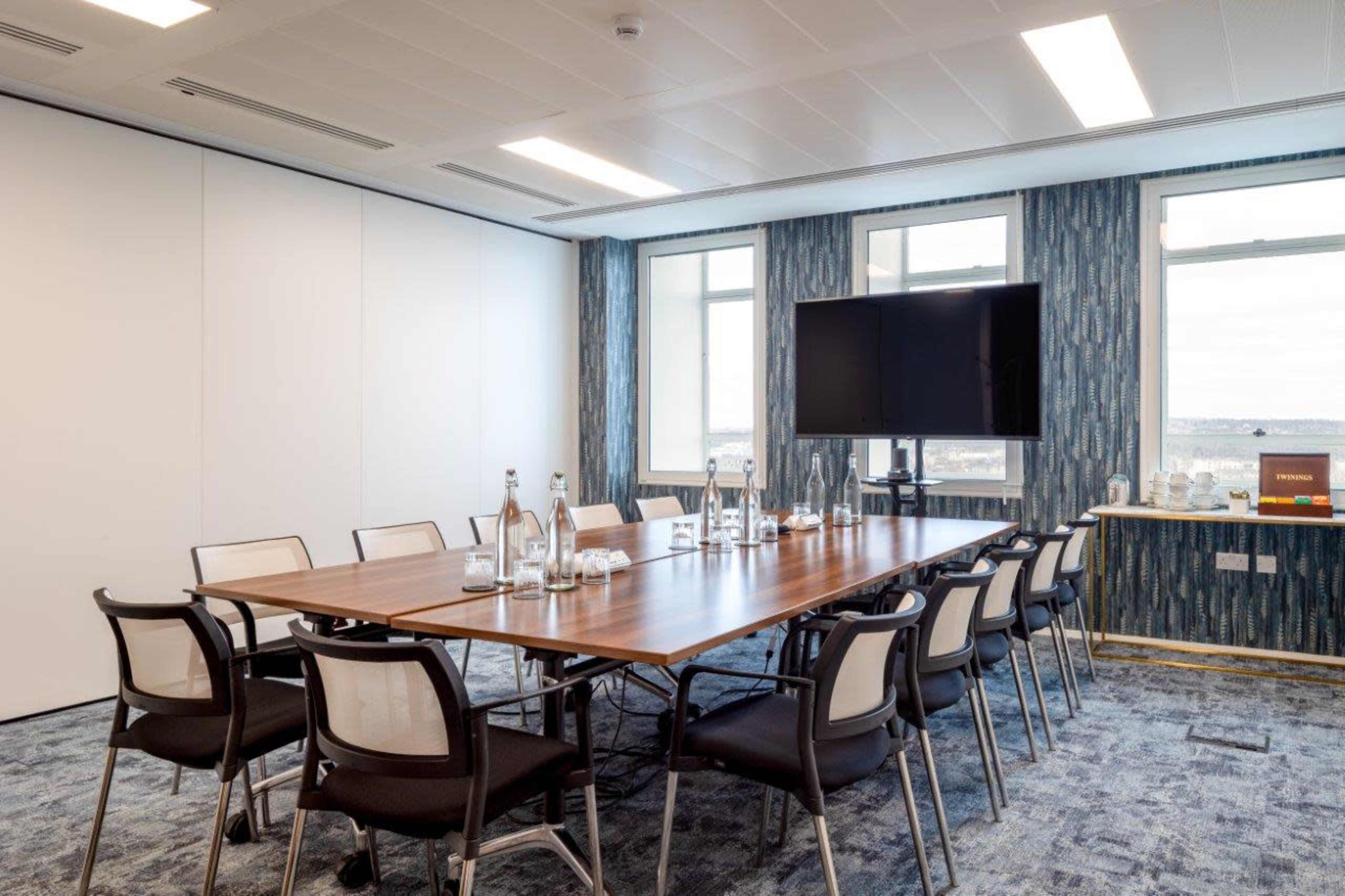 A conference room features a rectangular wooden table surrounded by chairs, with large windows and a television mounted on the wall.