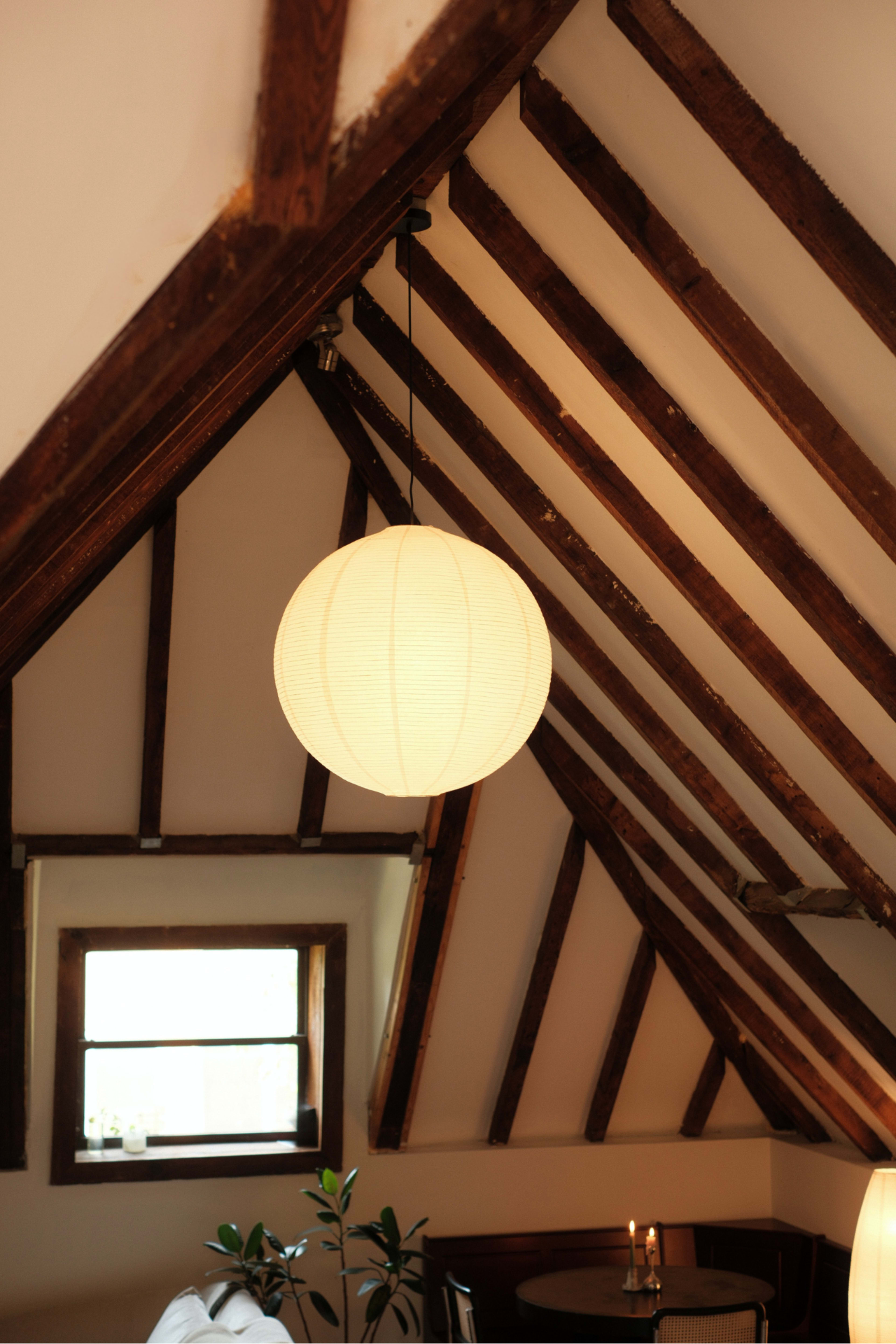 A round paper lantern hangs from the ceiling of a room with exposed wooden beams and a small window.