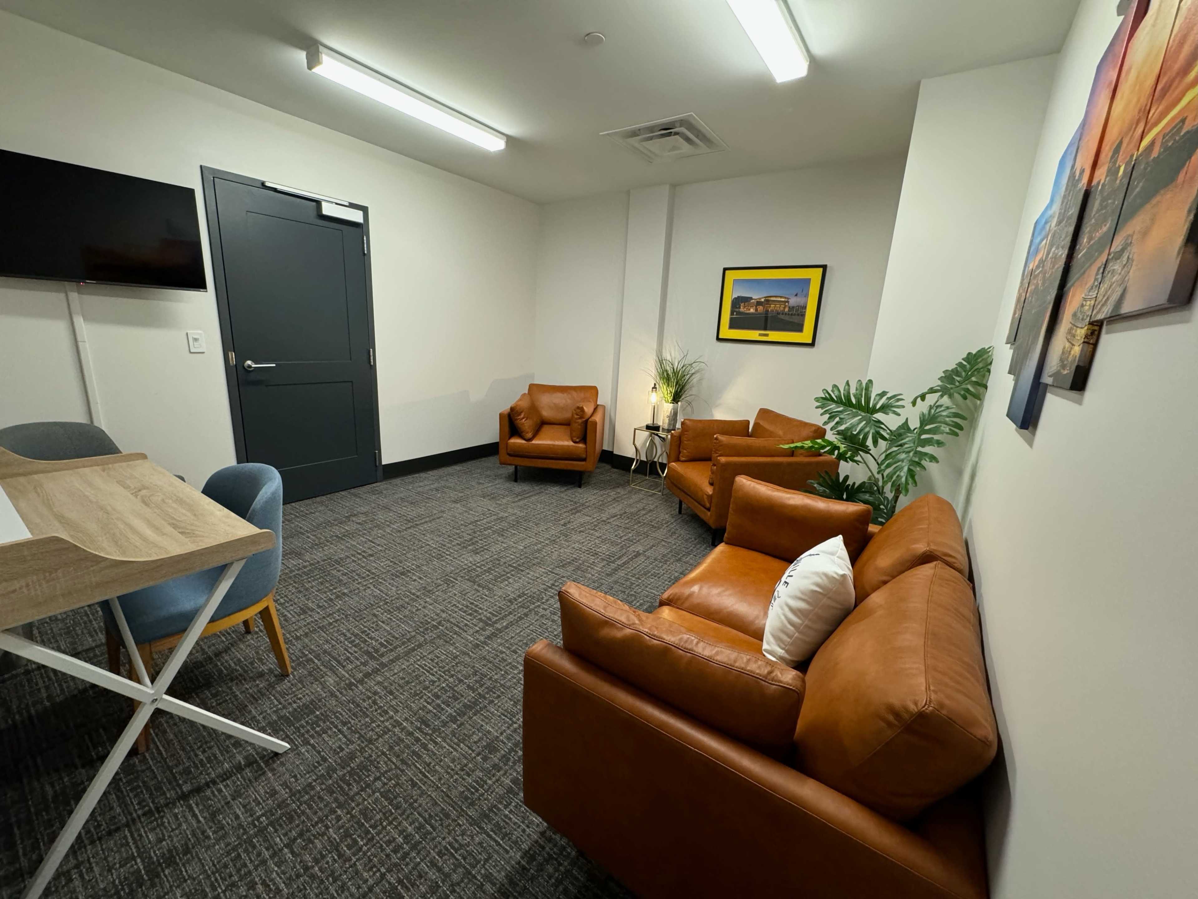 A small meeting room with two brown leather armchairs, a small table with a blue chair, a television on the wall, and a potted plant.