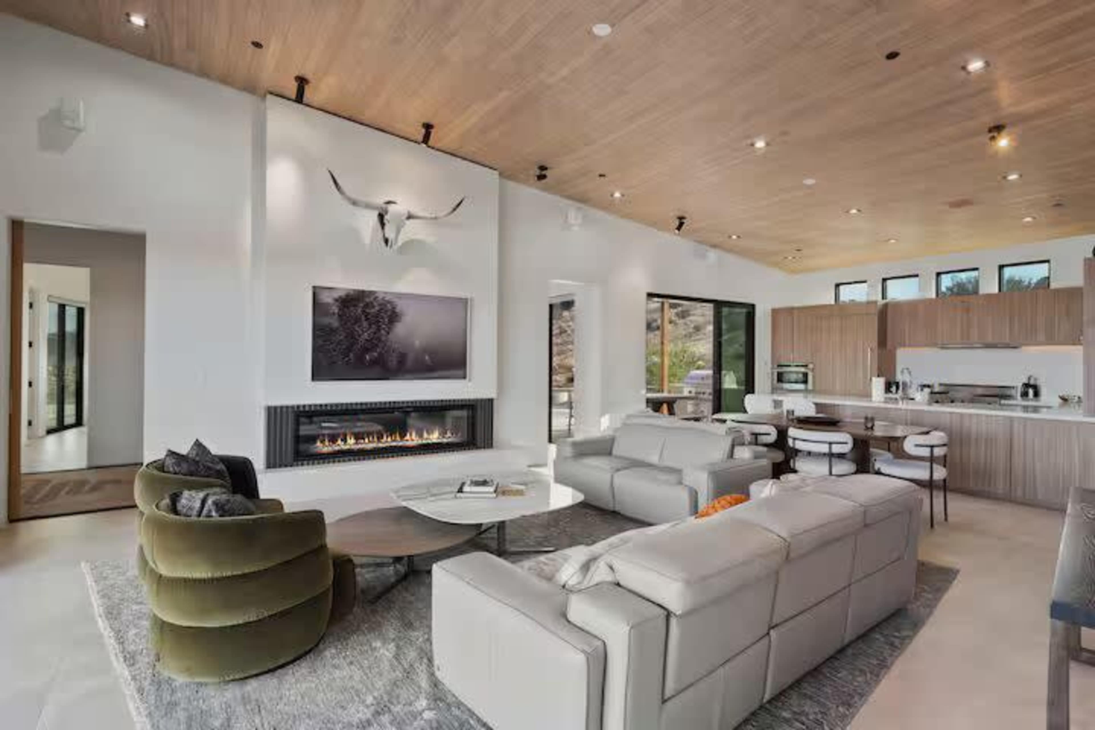 The image shows a modern living room featuring a fireplace, large windows, and an open kitchen area with sleek furniture.