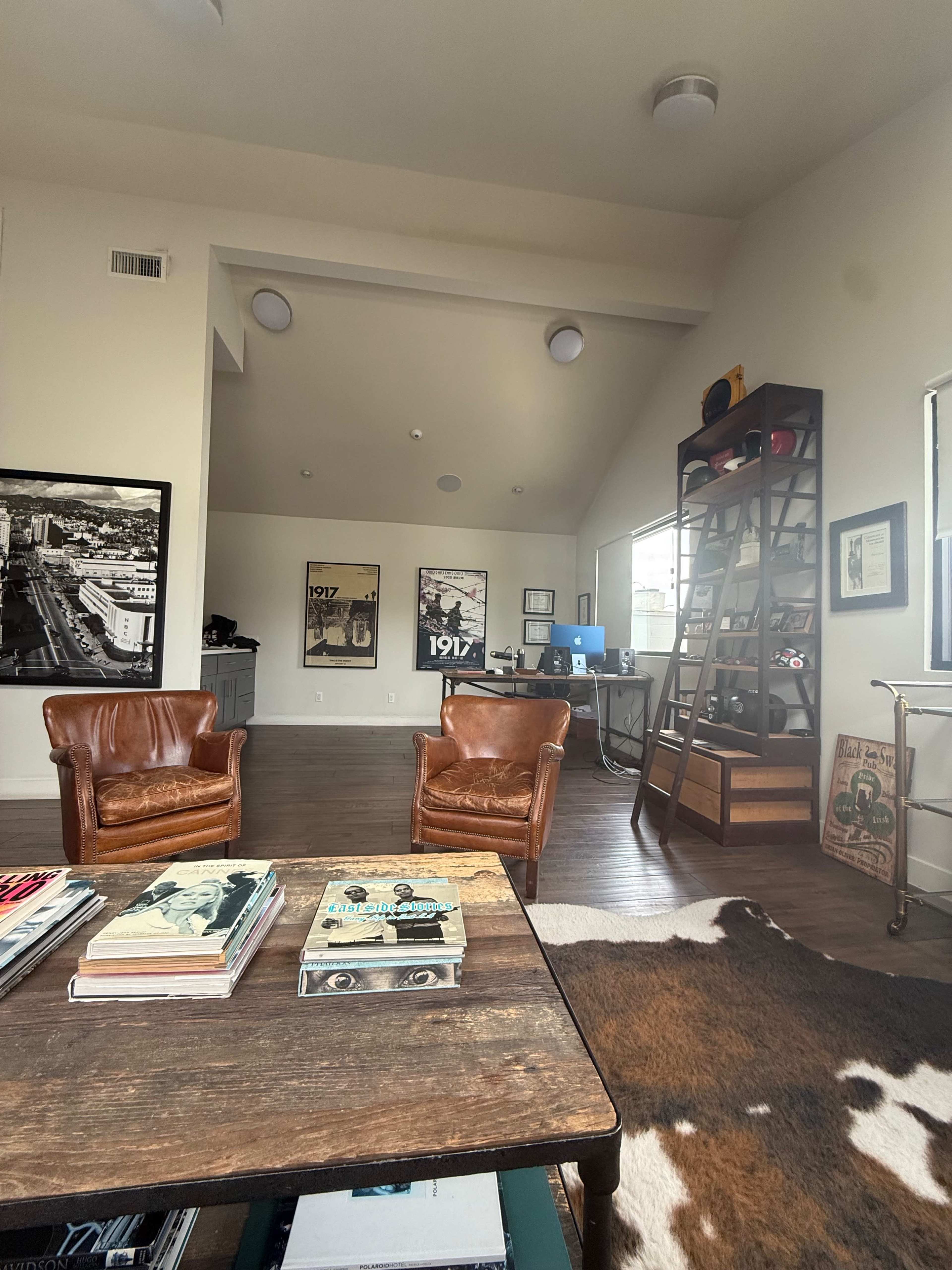The image depicts a modern office space featuring two brown leather chairs, a wooden coffee table with books, a bookshelf, and a desk with a computer in the background.
