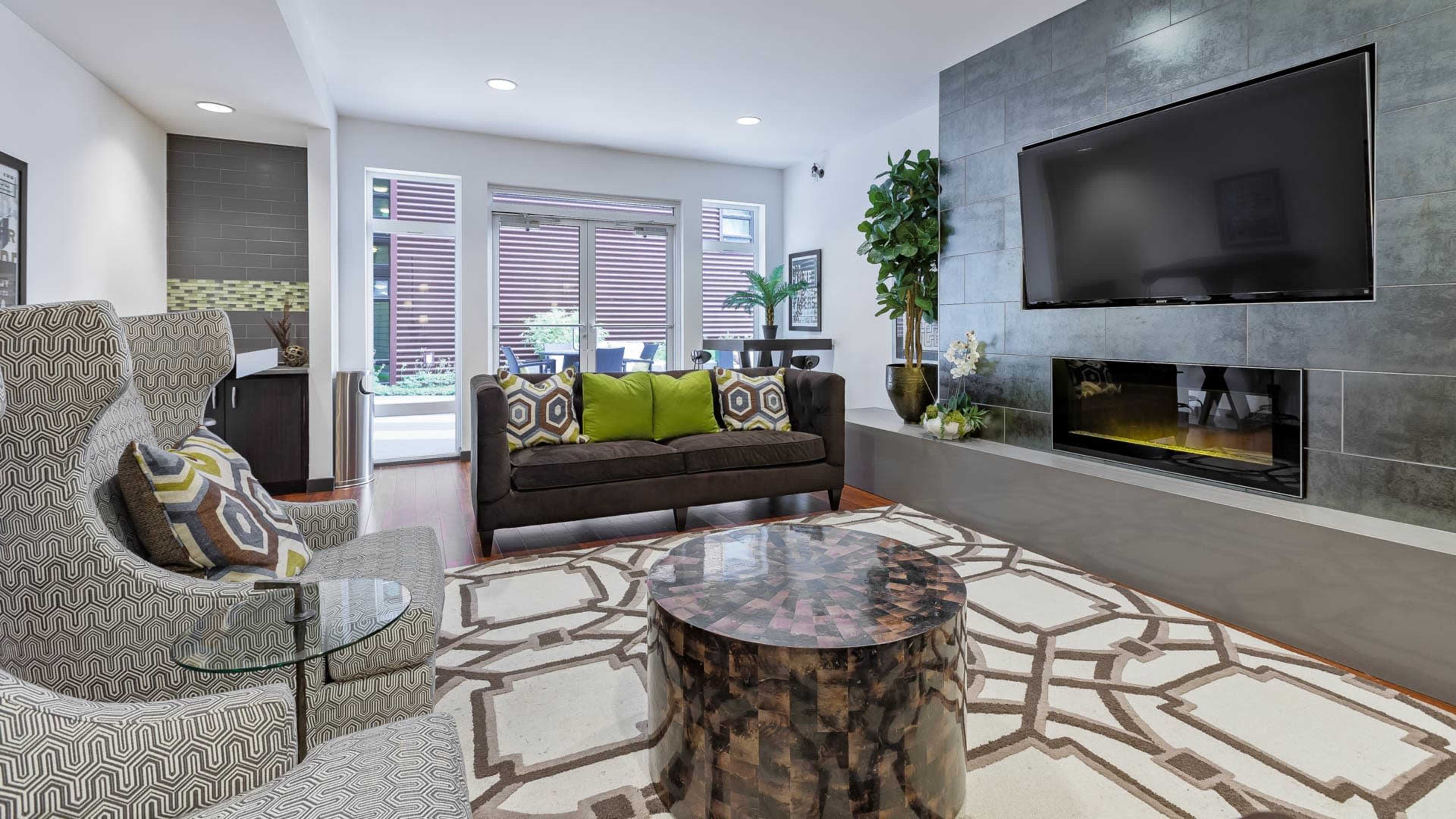 A modern living room features a patterned rug, a dark sofa with green and patterned pillows, two patterned chairs, a coffee table, a mounted television, and large windows with natural light.