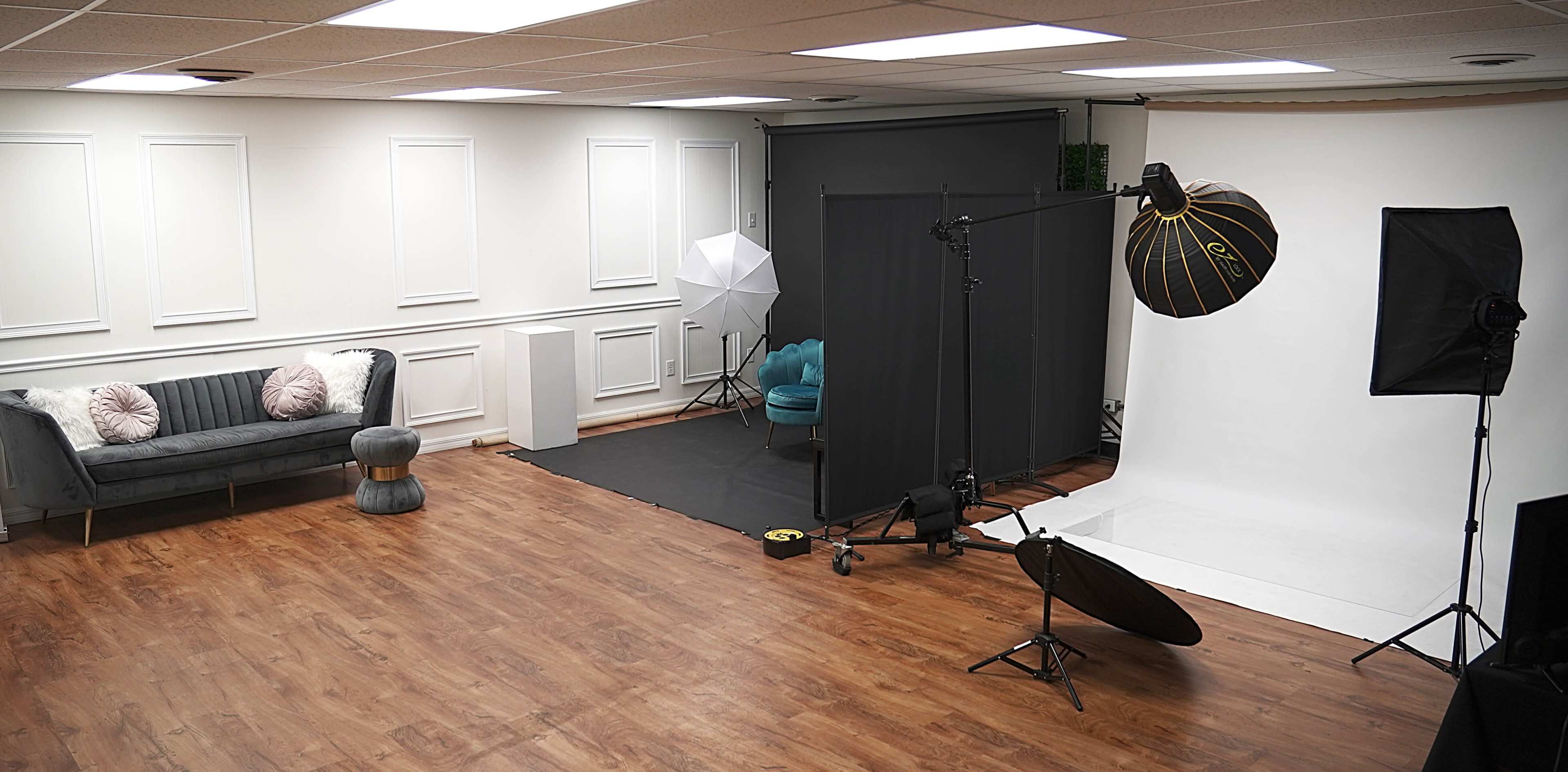 The image shows a photography studio with a gray sofa, lighting equipment, and a plain backdrop set up for shoots.