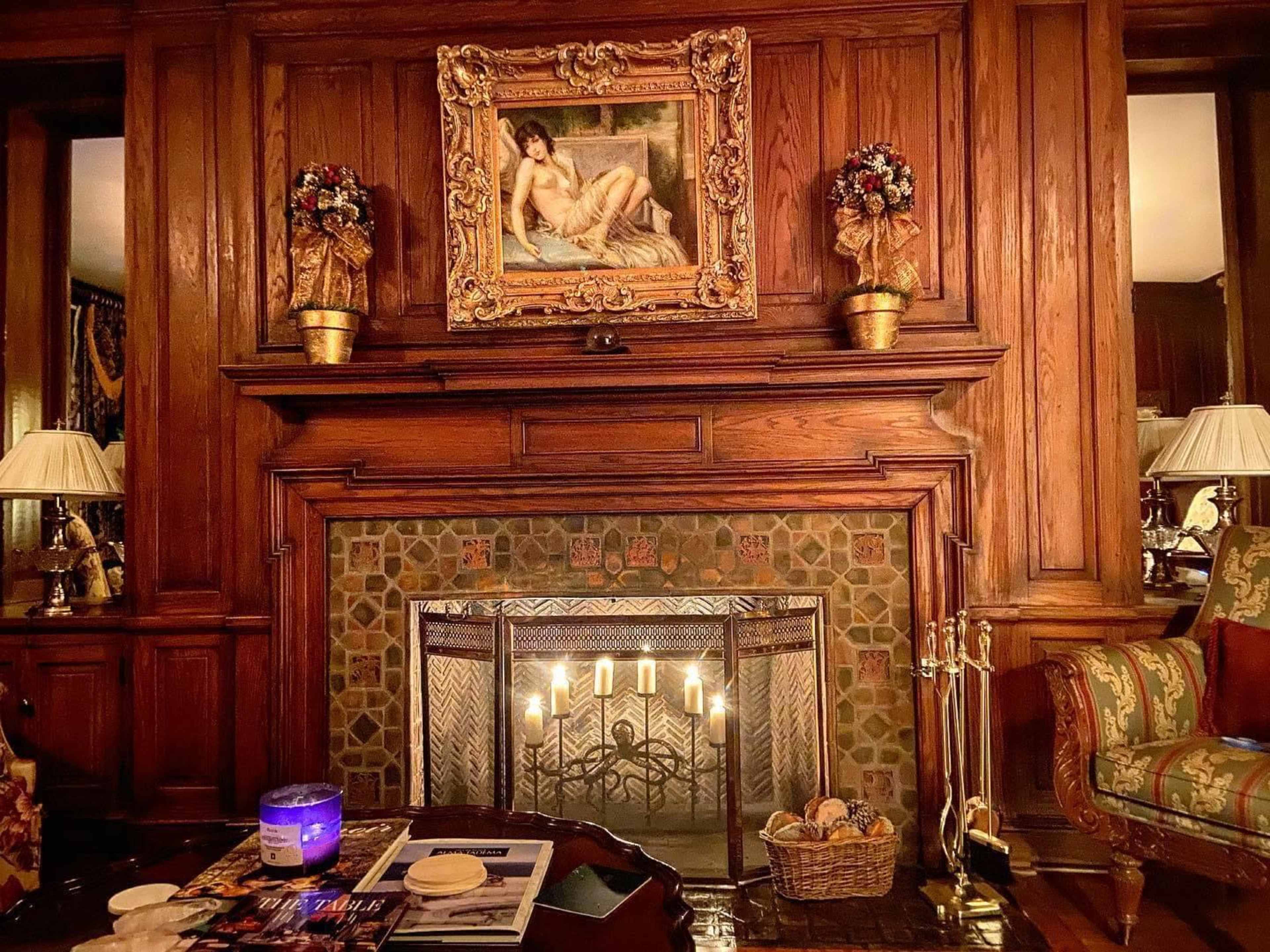 The image shows a wooden fireplace adorned with an ornate painting above it, surrounded by decorative vases and a cozy seating area.