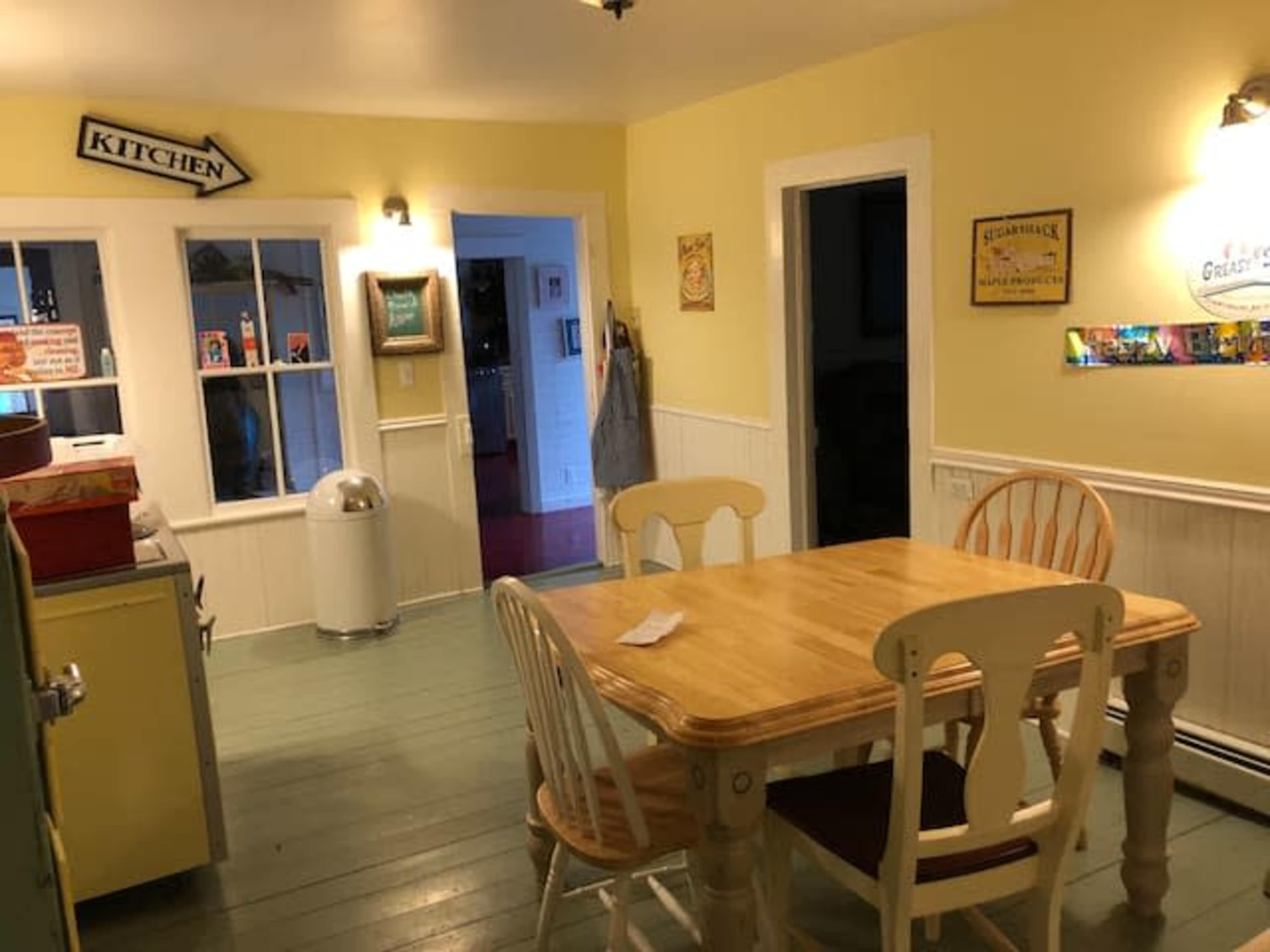 A brightly colored kitchen with a wooden table and chairs, a kitchen sign, and a door leading to another room.