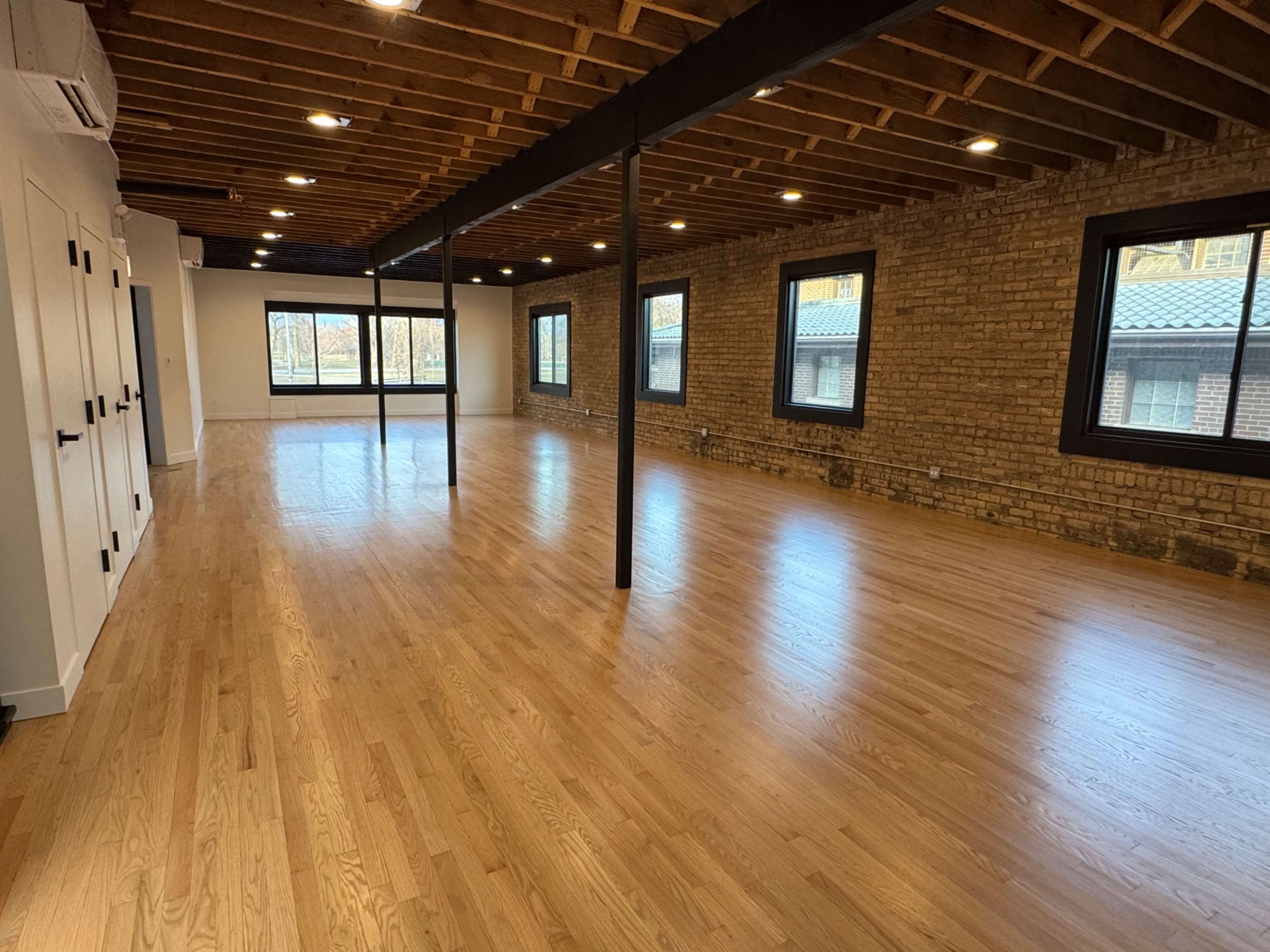 The image shows a spacious, empty room with hardwood flooring, exposed wooden beams, and large windows along the walls.