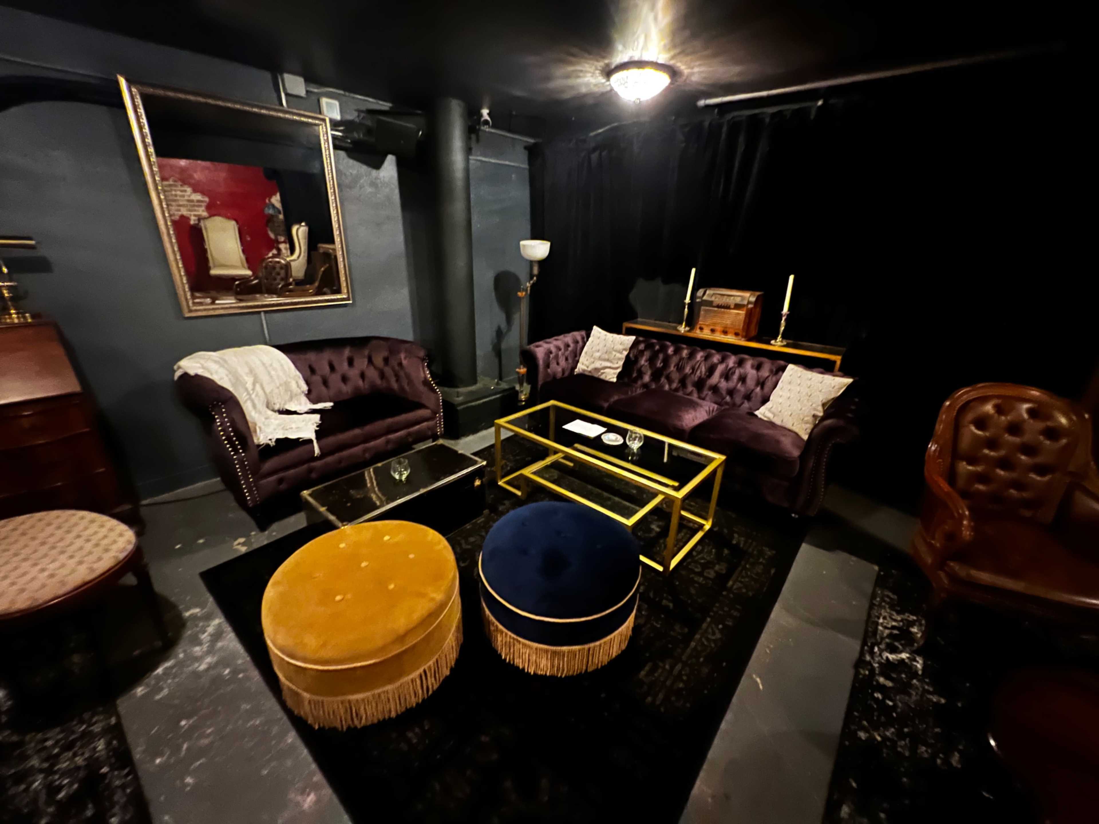A dimly lit lounge area featuring a pair of purple sofas, a glass coffee table, and two round ottomans, one blue and one yellow, set on a black rug.