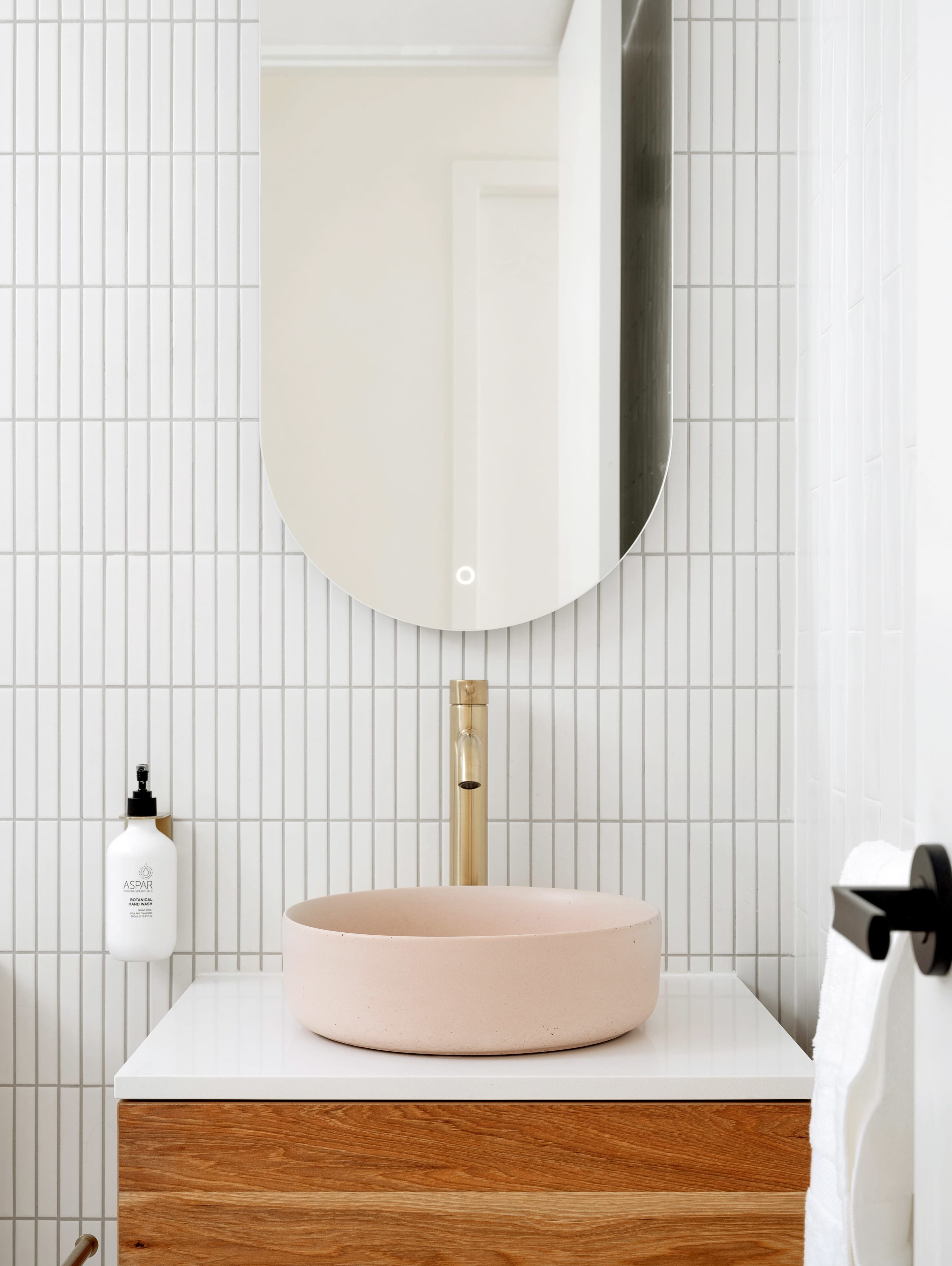 A modern bathroom features a pink ceramic sink mounted on a wooden cabinet, with a round mirror above and a soap dispenser on the side.