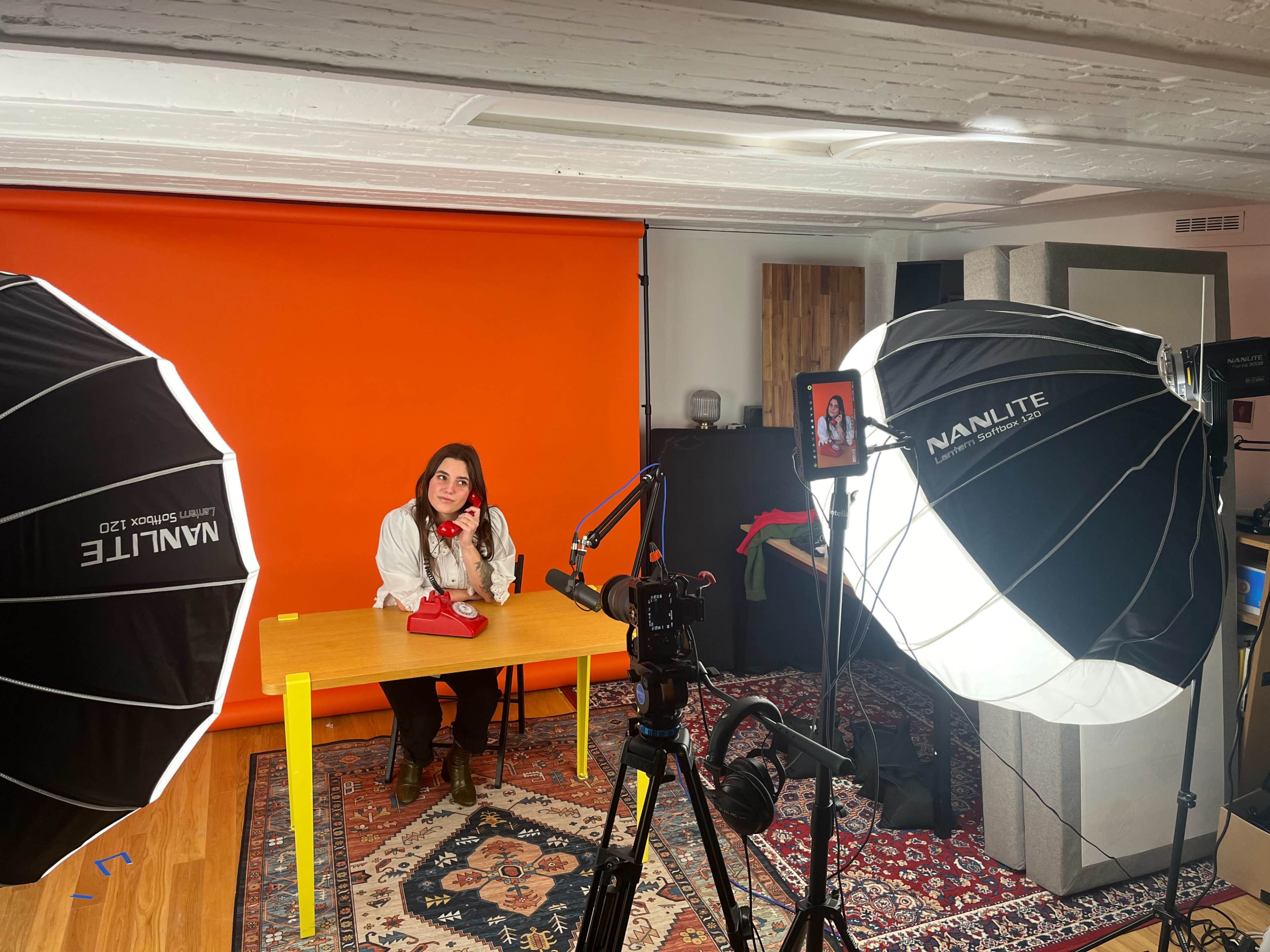 A woman sits at a table with a red purse in front of an orange backdrop, flanked by large softbox lights and a camera setup.