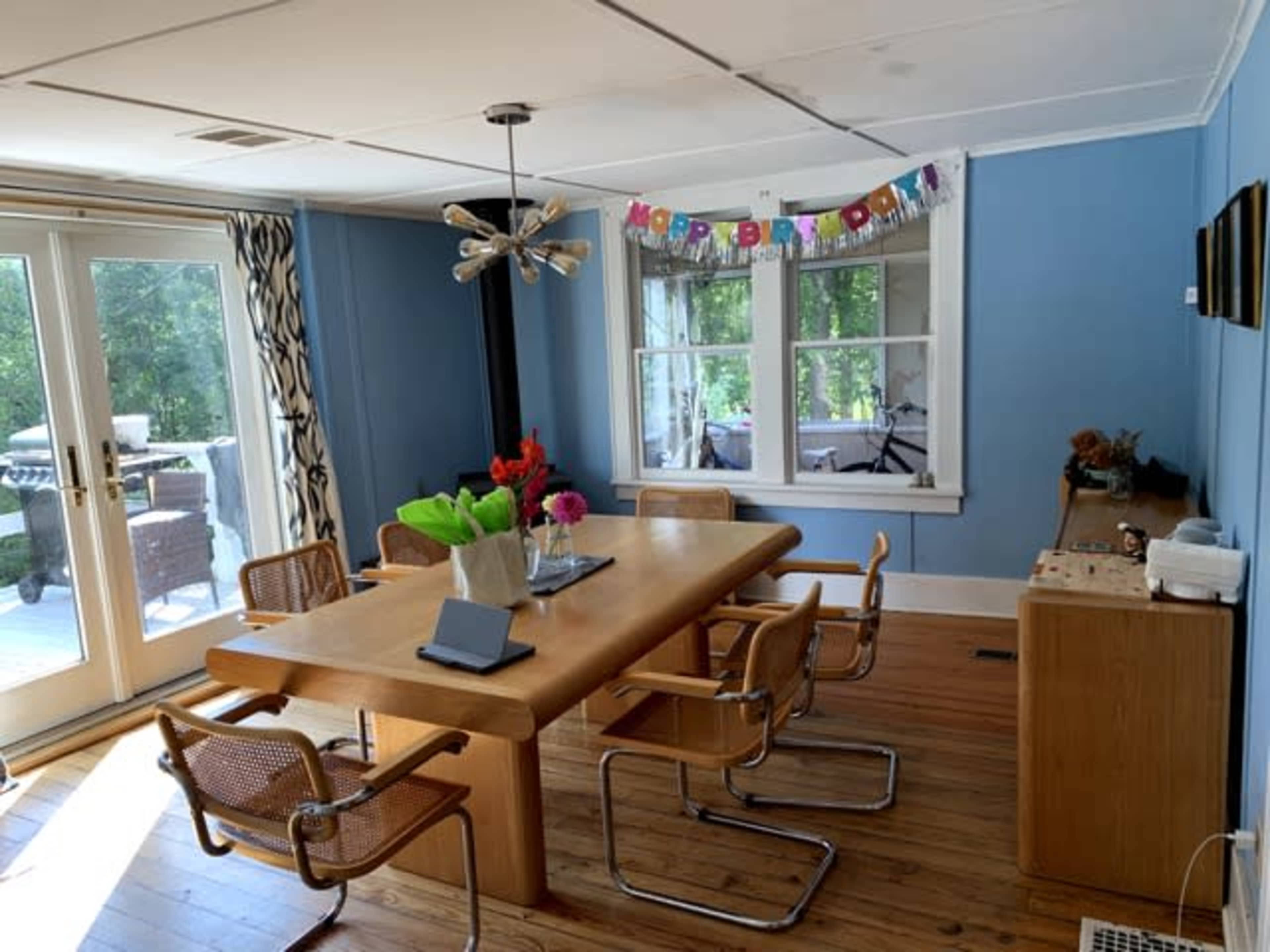 A dining area with a wooden table surrounded by modern chairs, a birthday banner displayed on the wall, and a view of the outdoor greenery through large windows.