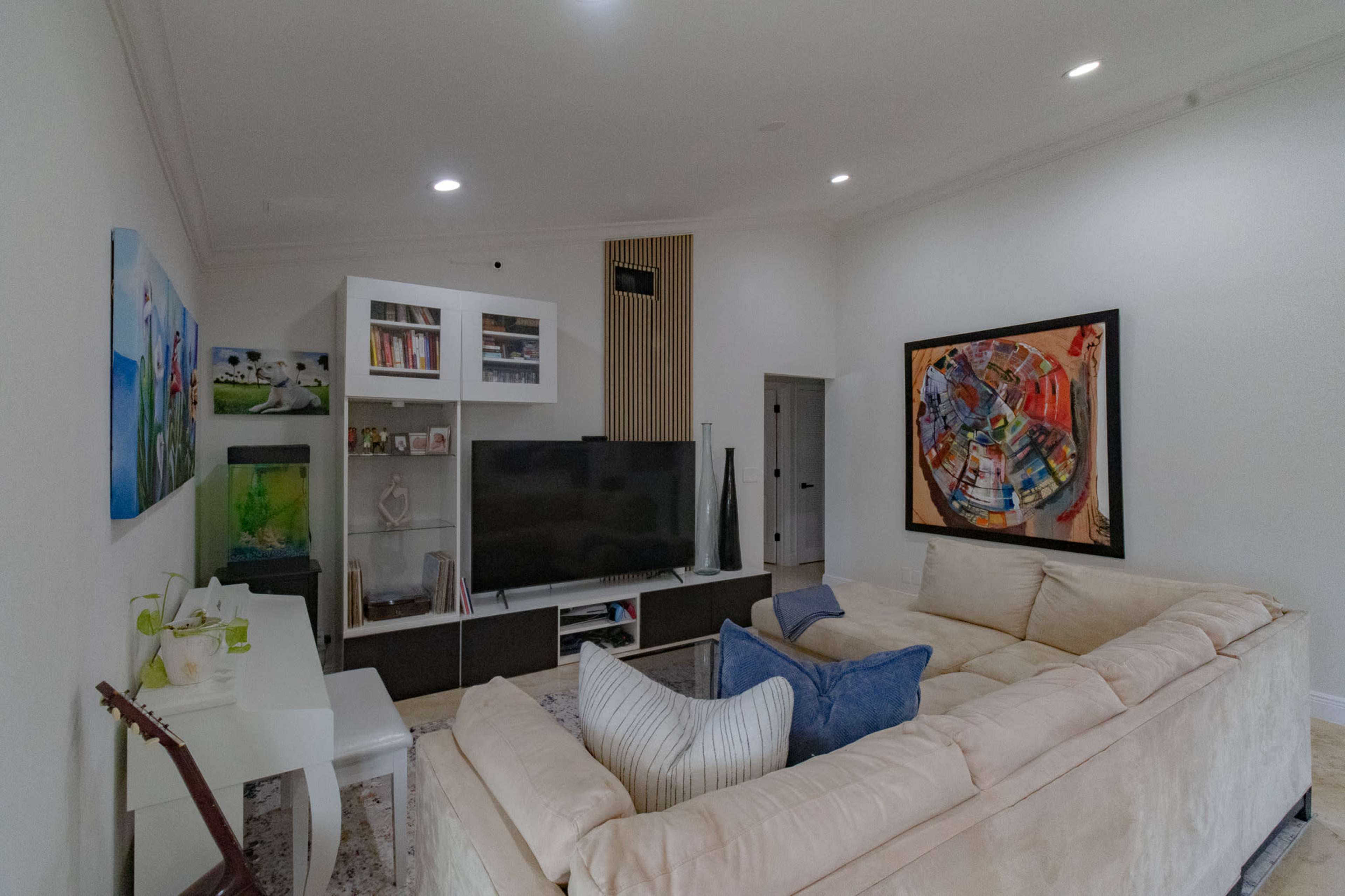 The living room features a beige sectional sofa, a large flat-screen TV mounted on a wall unit, and a mix of artwork and decorative items on display.