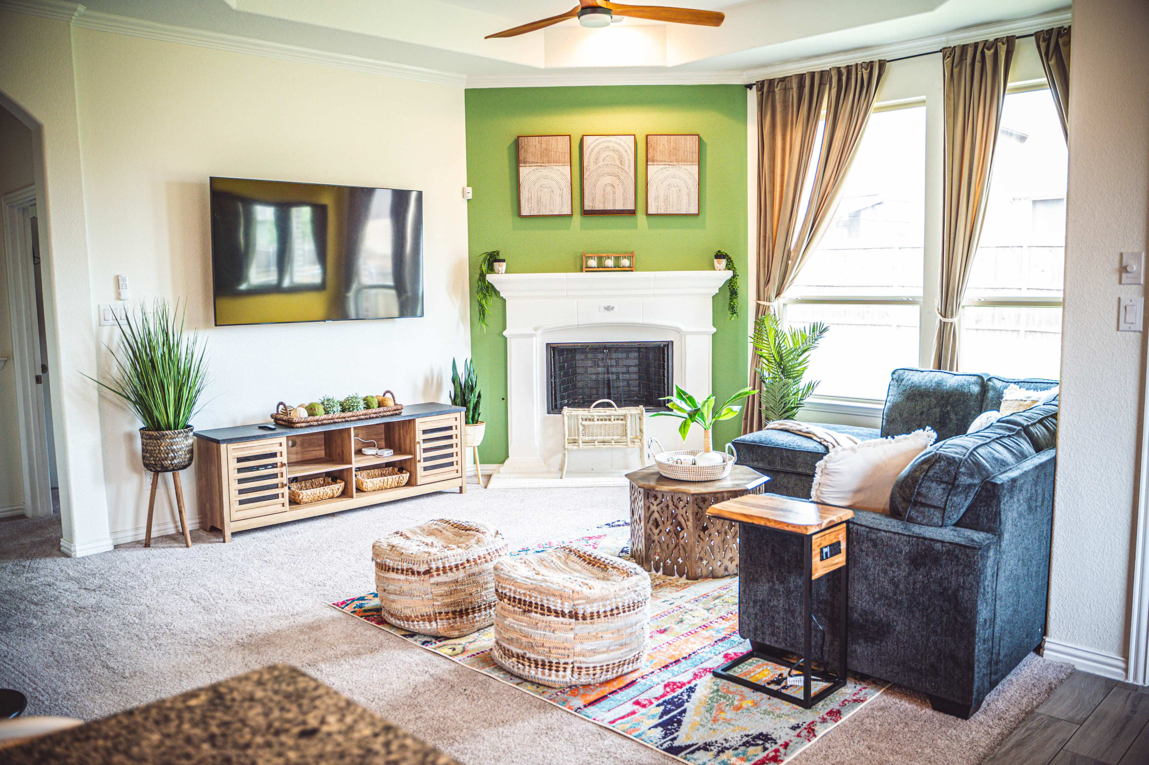 A cozy living room with a large television on a cream-colored wall, a green accent wall, a fireplace, and a seating area featuring a dark sofa and decorative poufs.