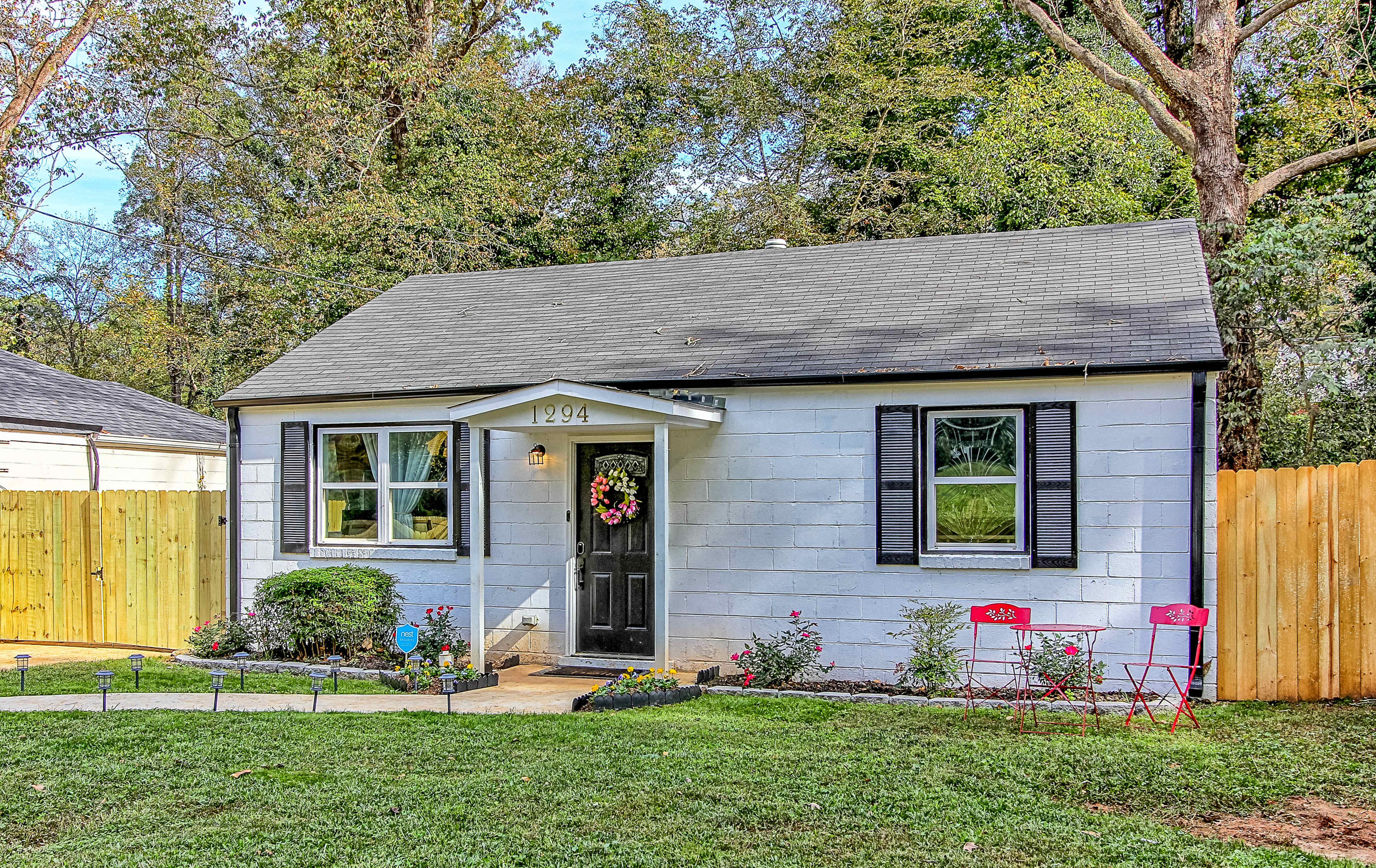 A small, single-story house with light blue siding and black shutters features a front porch with a decorative wreath and two red chairs on the lawn.