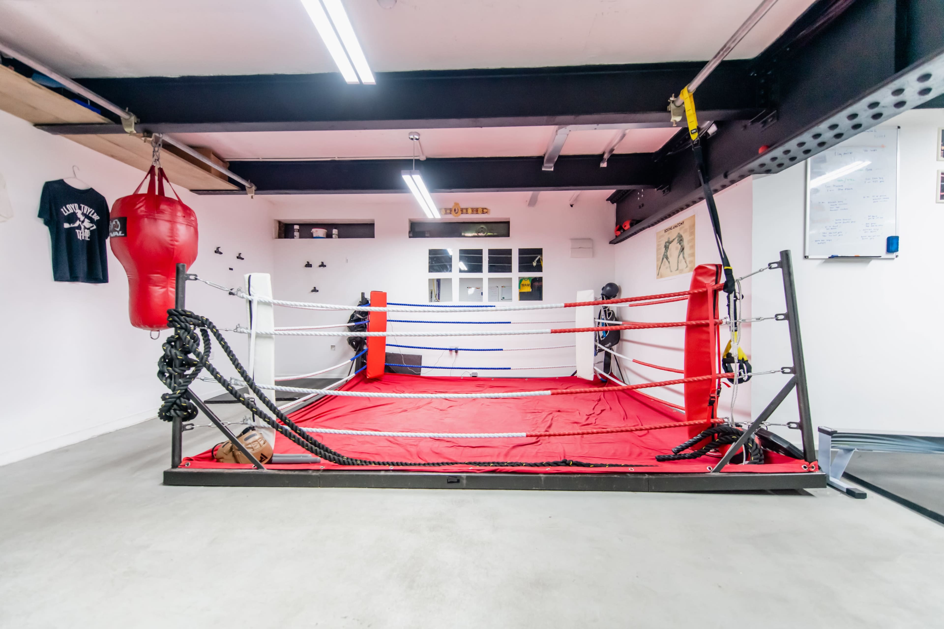 A boxing ring in a gym with red and white ropes, a heavy bag hanging beside it, and various training equipment on the walls.